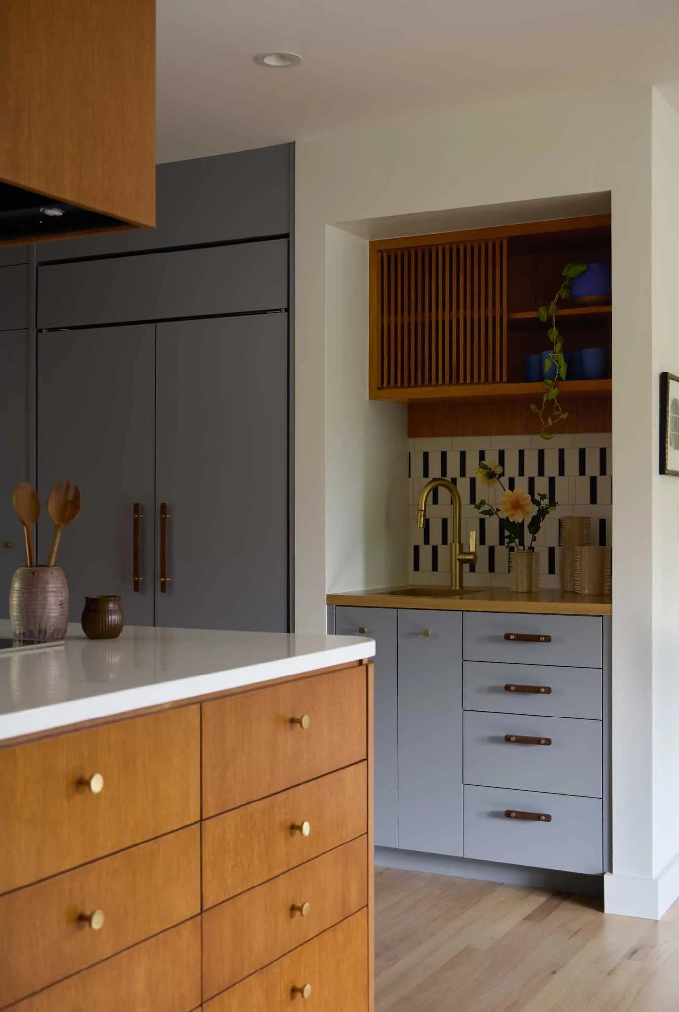 A modern kitchen with a white countertop, wooden and gray cabinets, a gold faucet, and a backsplash with black and white tiles. There are vases with flowers and kitchen utensils on the counter.