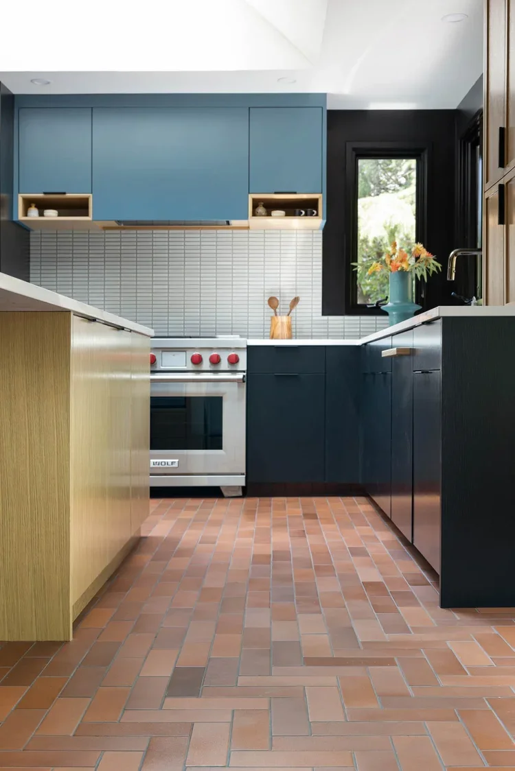 Modern kitchen with blue upper cabinets, dark lower cabinets, a white brick backsplash, and terracotta tile flooring. A window with a view of greenery, a vase with flowers, and a wooden utensil holder are visible.
