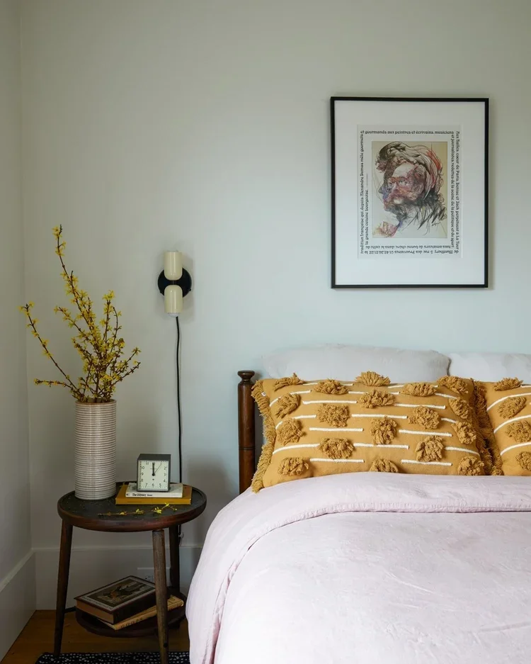 Bedroom with a nightstand, vase with yellow flowers, wall-mounted light fixture, framed artwork of a woman's profile, and a bed with decorative yellow pillows.