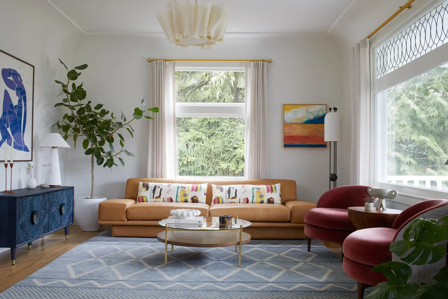 Living room with a tan sofa, red armchairs, blue sideboard, large potted plant, and windows with white curtains showing a green outdoor scene.