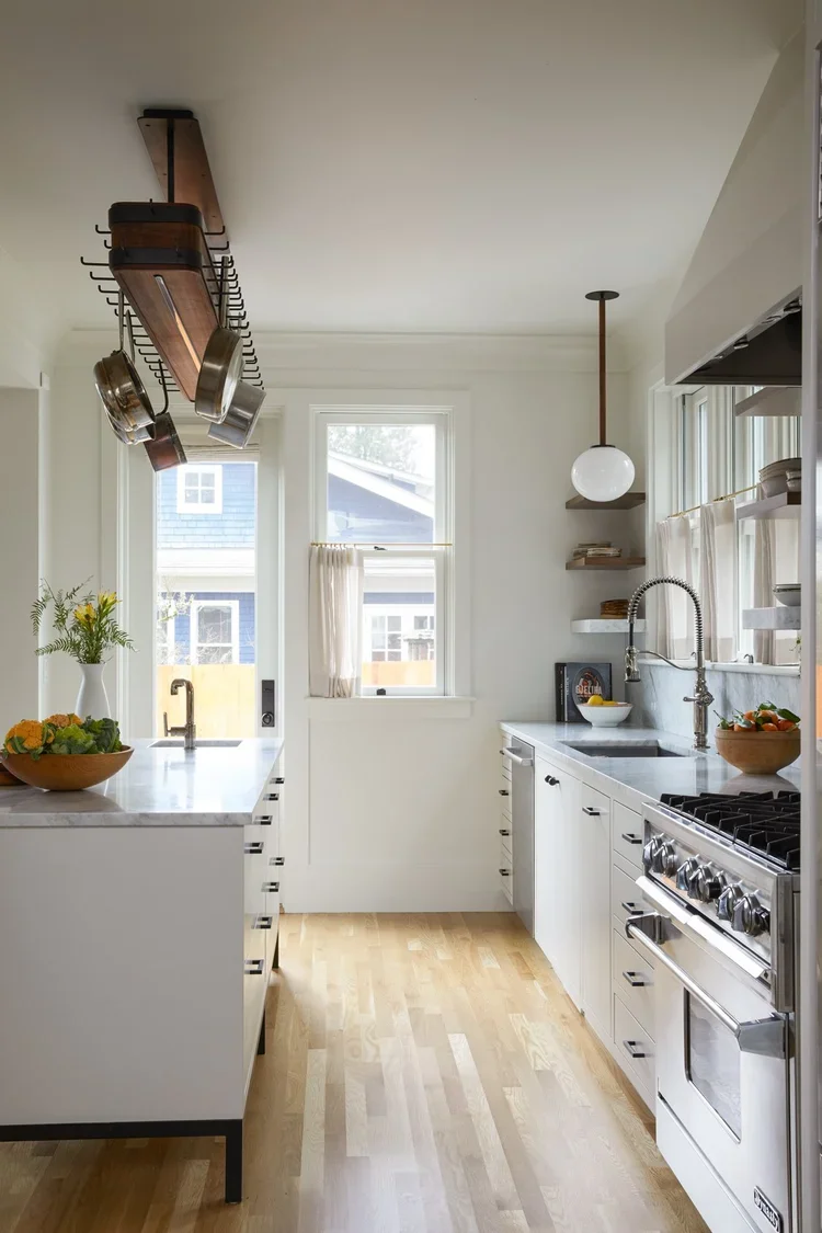 Bright kitchen with white cabinets, wooden floors, and natural light from windows, featuring pots and bowls on the countertops and hanging cookware