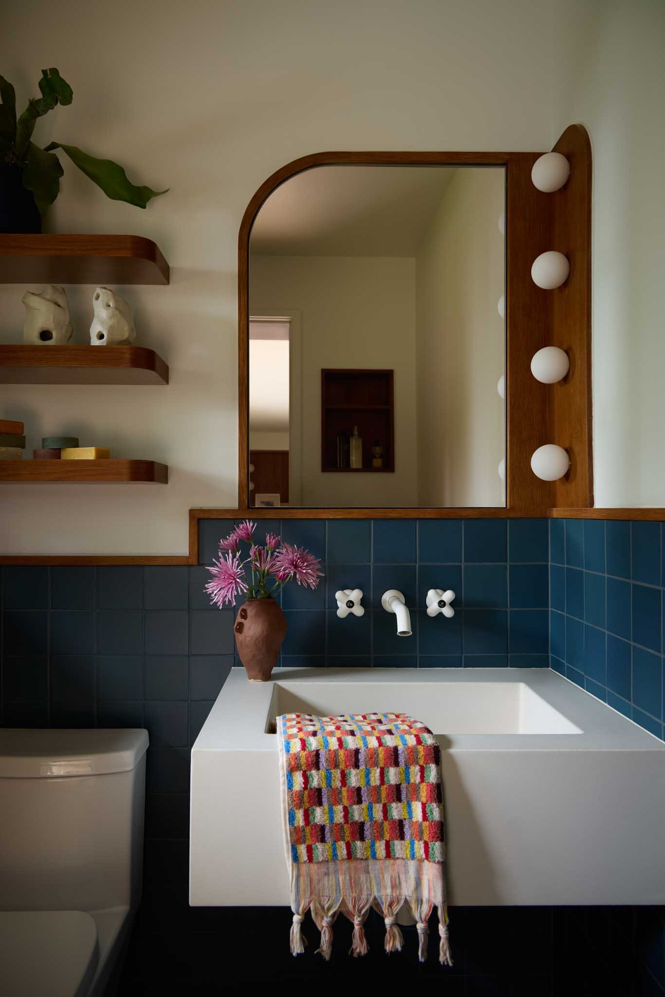 A bathroom with a white sink, a colorful striped towel, blue tiles, a brown vase with pink flowers, a mirror with wooden frame and round lights, and wooden shelves with decorative items.