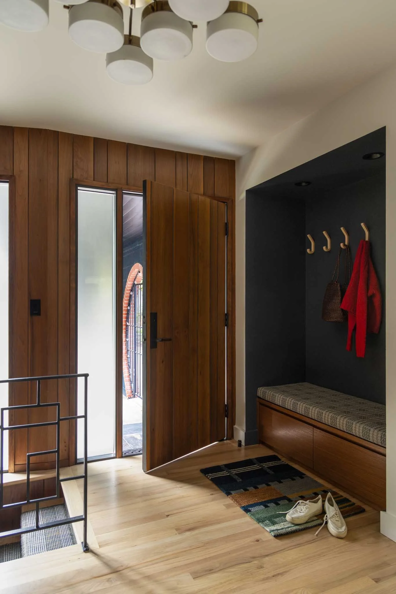 Entryway with wooden door, shoes on the floor, hooks with red coat, woven bench, and a black wall background.