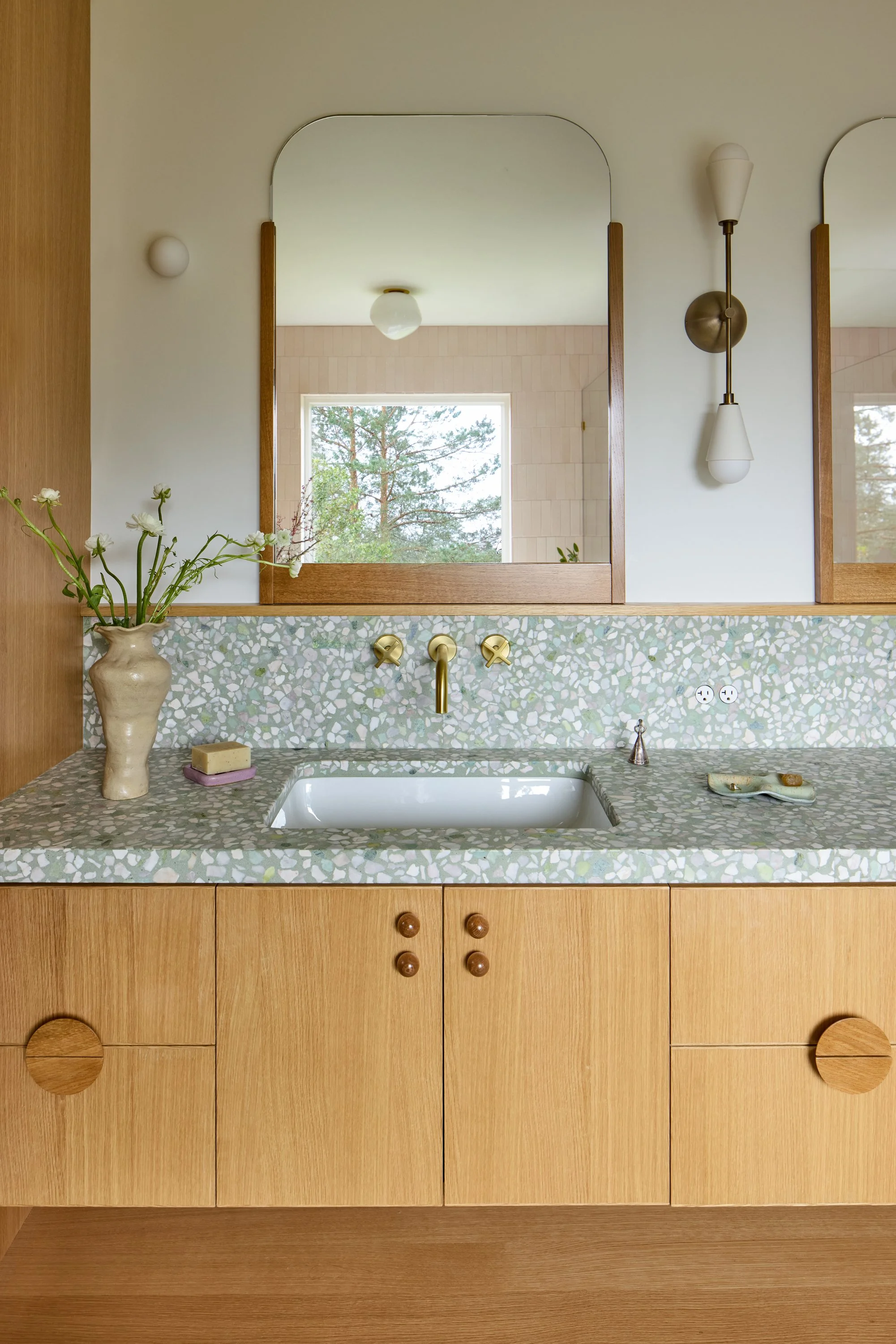 A bathroom vanity with a terrazzo countertop, wooden cabinetry, a vase with flowers, and a mirror reflecting a window and outdoor trees.