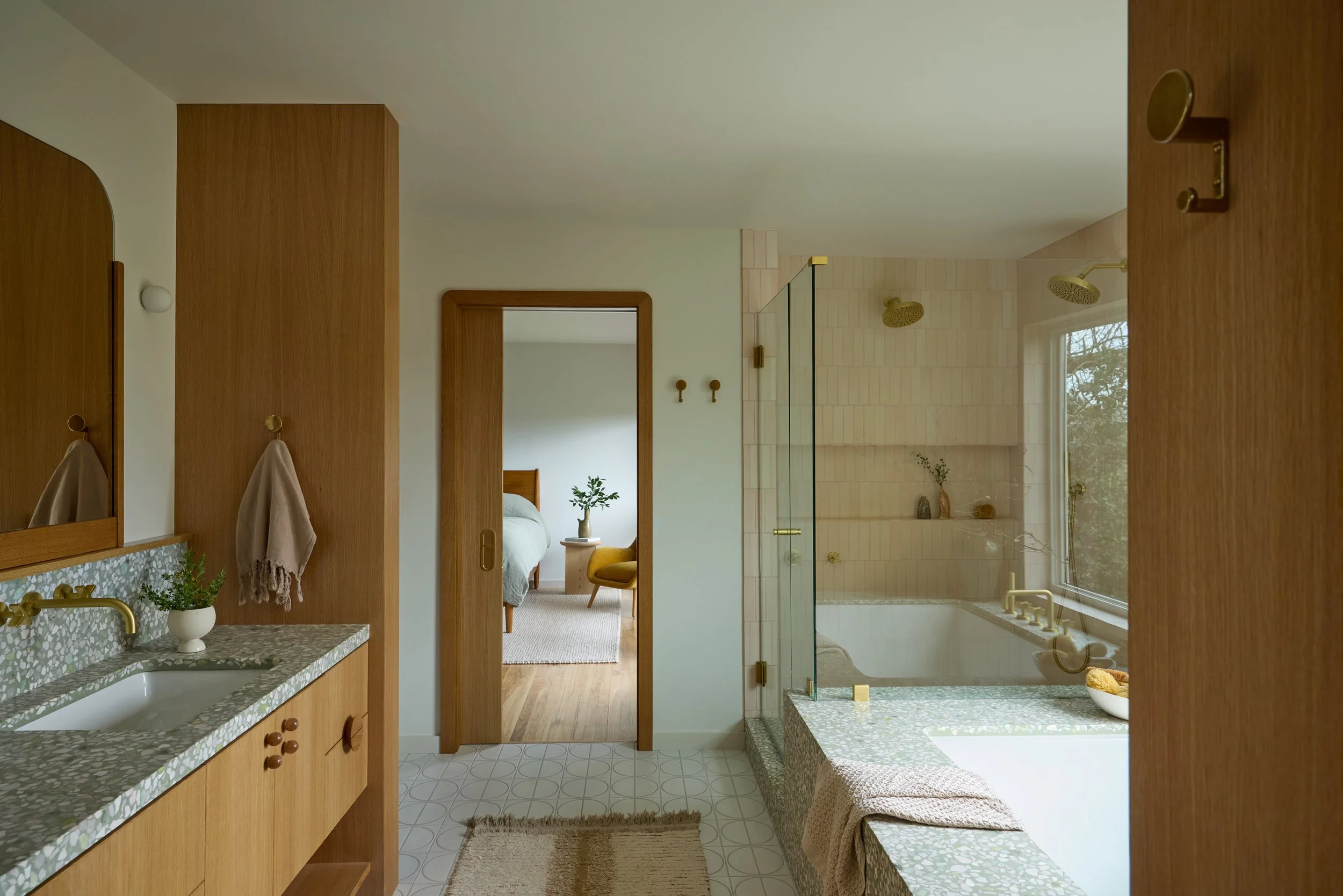 A modern bathroom with wood accents, a shower with a glass door and gold fixtures, and a view into a bedroom with a bed, a yellow chair, and a small table with a plant.