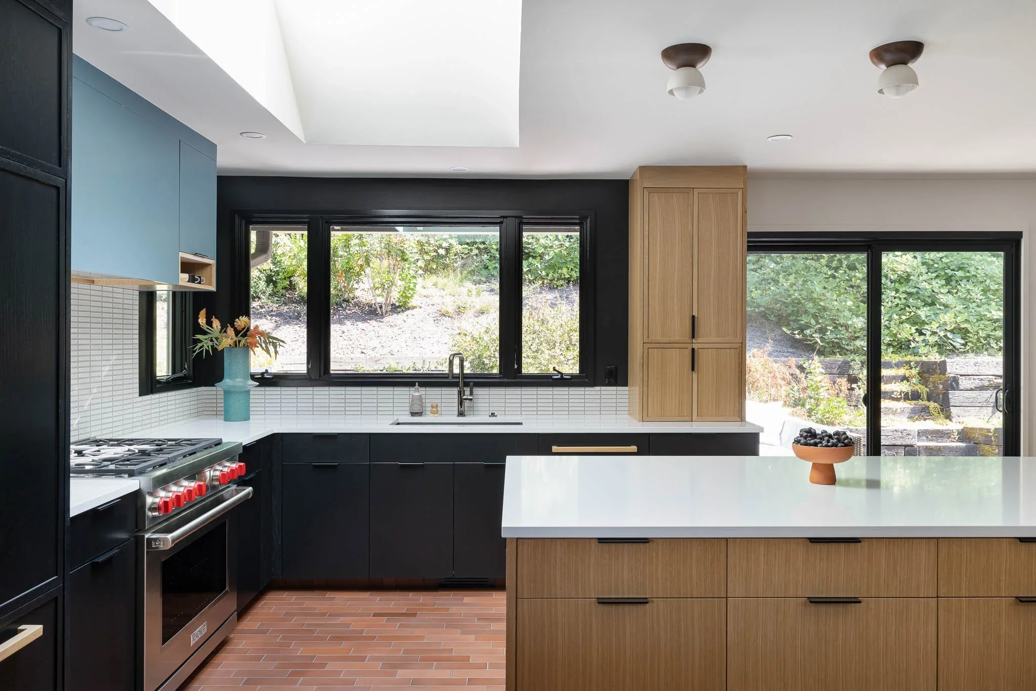 Modern kitchen with black and wood cabinetry, white countertops, a window above the sink, and a sliding glass door leading outside.