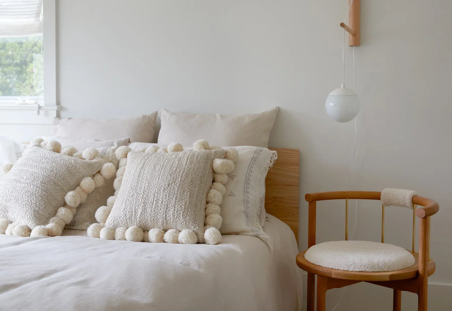 A cozy bedroom with a bed decorated with white and cream pillows, a small wooden chair with a cushioned seat, and a modern hanging light fixture on the wall.
