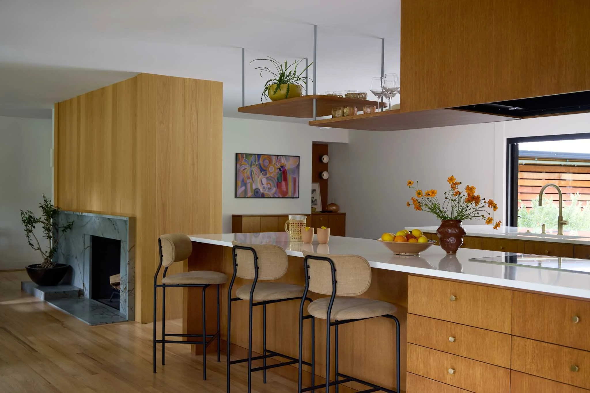 Modern kitchen with wooden cabinetry, a white island with chairs, a flower arrangement, and a view of the outdoors through a window.