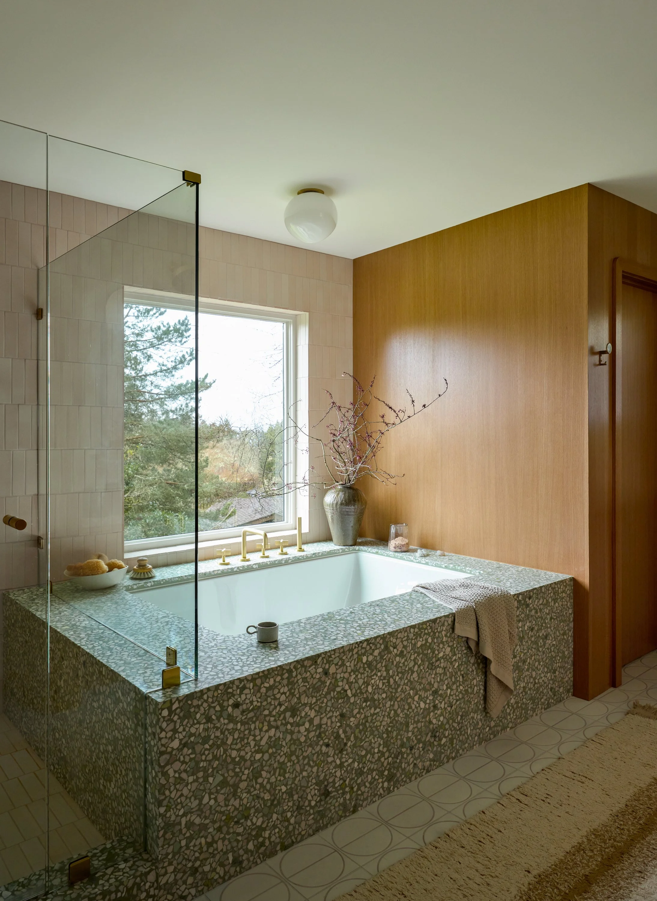 View of a bathroom with a large built-in bathtub surrounded by green and white mosaic tiles. The bathtub area has a window overlooking trees, a dried branch arrangement in a vase, and various bath items, with a towel hanging over the edge. The ceiling is white, and one wall is covered with wooden paneling, with a section of beige tiled wall and a glass shower partition.