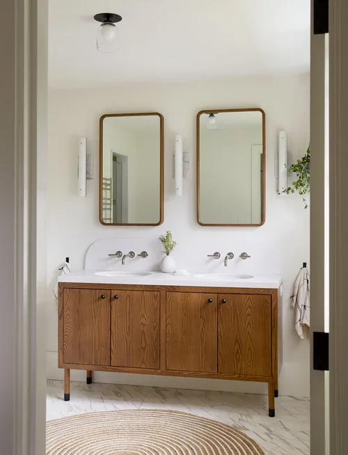 Bathroom with two mirrors above a wooden vanity, white walls, and minimal decor including a small vase with greenery and a hanging potted plant.