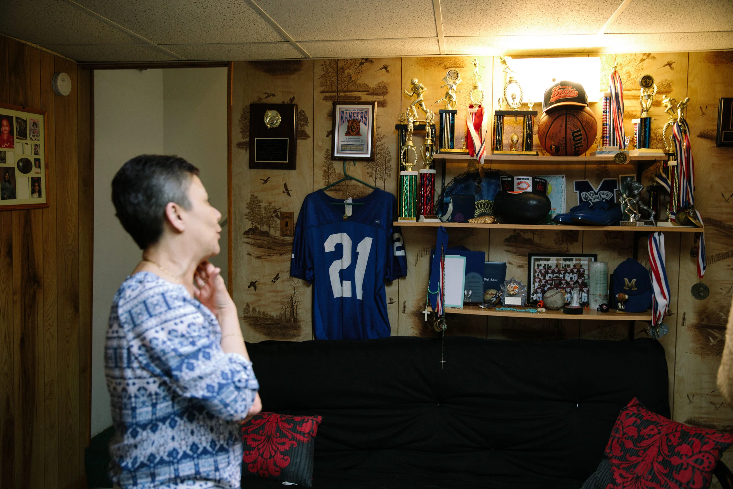 Elizabeth Ramirez, stands in the basement of her home gazing at a wall of rememberence devoted to her late son Dee Jay. The shelves contain momentos of Dee Jay's life and death, with items like his high school diploma, sports trophies, and the program from his memorial service. Dee Jay was shot and killed in the Belmont-Cragin neighborhood of Chicago in 2011. Dee Jay's mother, Elizabeth, helped found an organization called Parents for Peace and Justice to help provide support and advocacy for families who have also lost children to gun violence in Chicago. MCC has partnered with Parents for Peace and Justice to help support them in their important work.