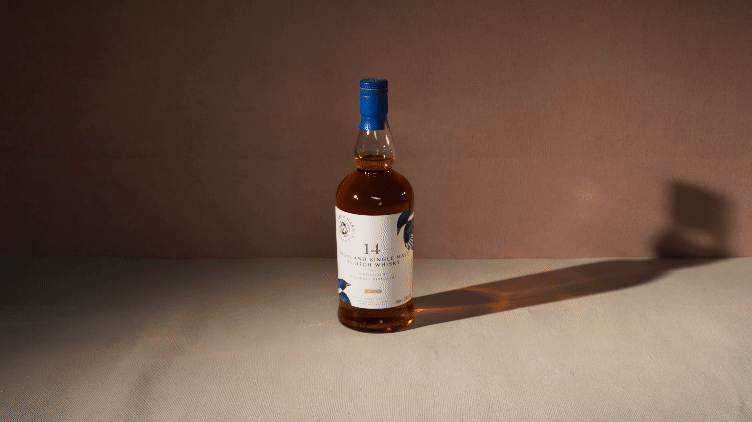 A small bottle of whiskey with a blue cap standing on a white surface, casting a shadow to the right, against a plain beige background.