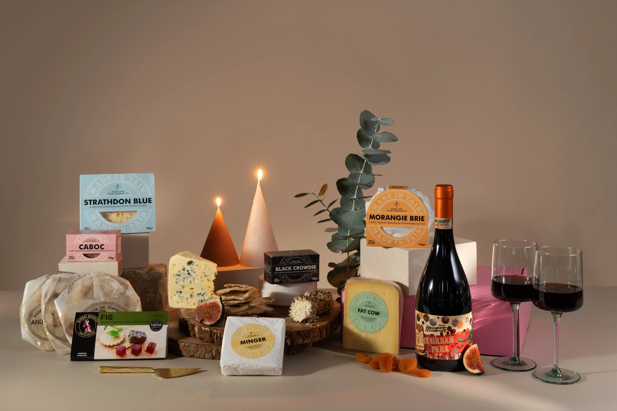 Cheese platter with various cheeses, crackers, figs, and garnishes, two glasses of red wine, decorative candles, eucalyptus branch, and a pink gift box in the background.