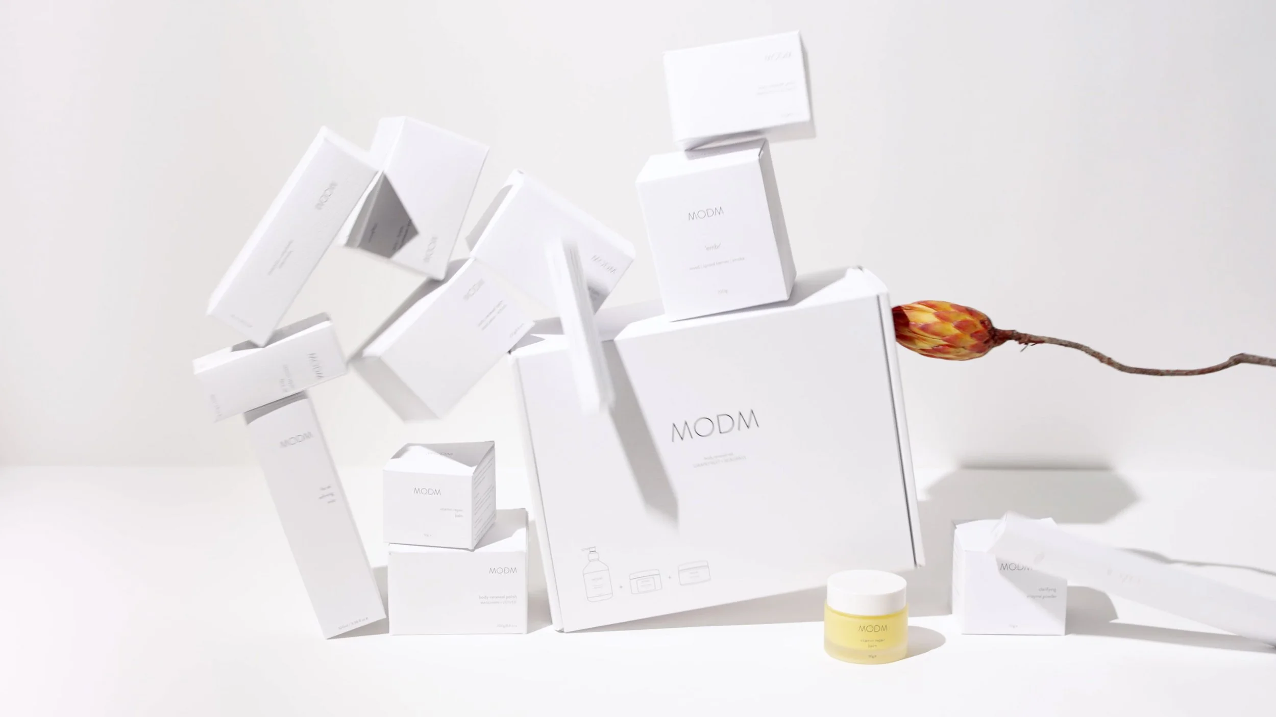 A collection of white skincare product boxes and a small yellow jar with a white lid, arranged against a white background, with a decorative flower stem.