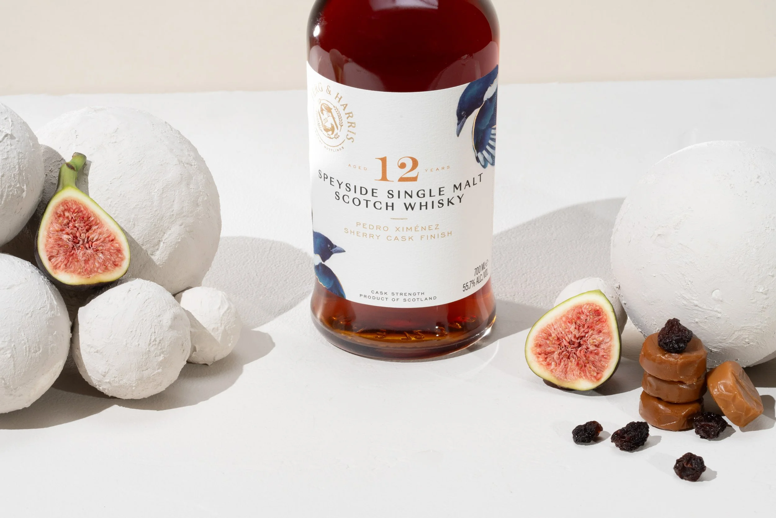 A bottle of 12-year-old Speyside single malt Scotch whisky with a white label featuring a bird design, surrounded by halved fresh figs, white textured spherical objects, and caramel candies with dried fruit on a white background.