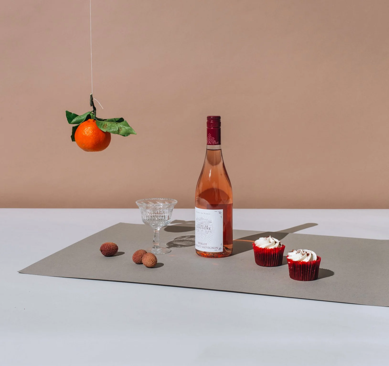 A still life arrangement with a hanging orange with green leaves, a pink wine bottle, a clear glass, three lychee fruits, and two red cupcakes with white frosting on a beige mat on a white surface.
