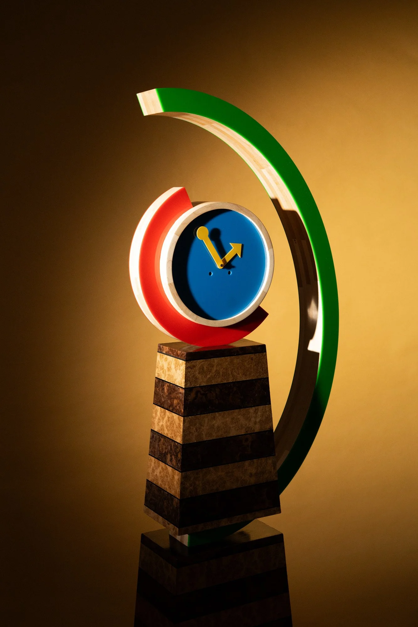 Colorful modern clock sculpture with a circular face, yellow clock hands, red and green curved accents, mounted on a striped black and brown pedestal.