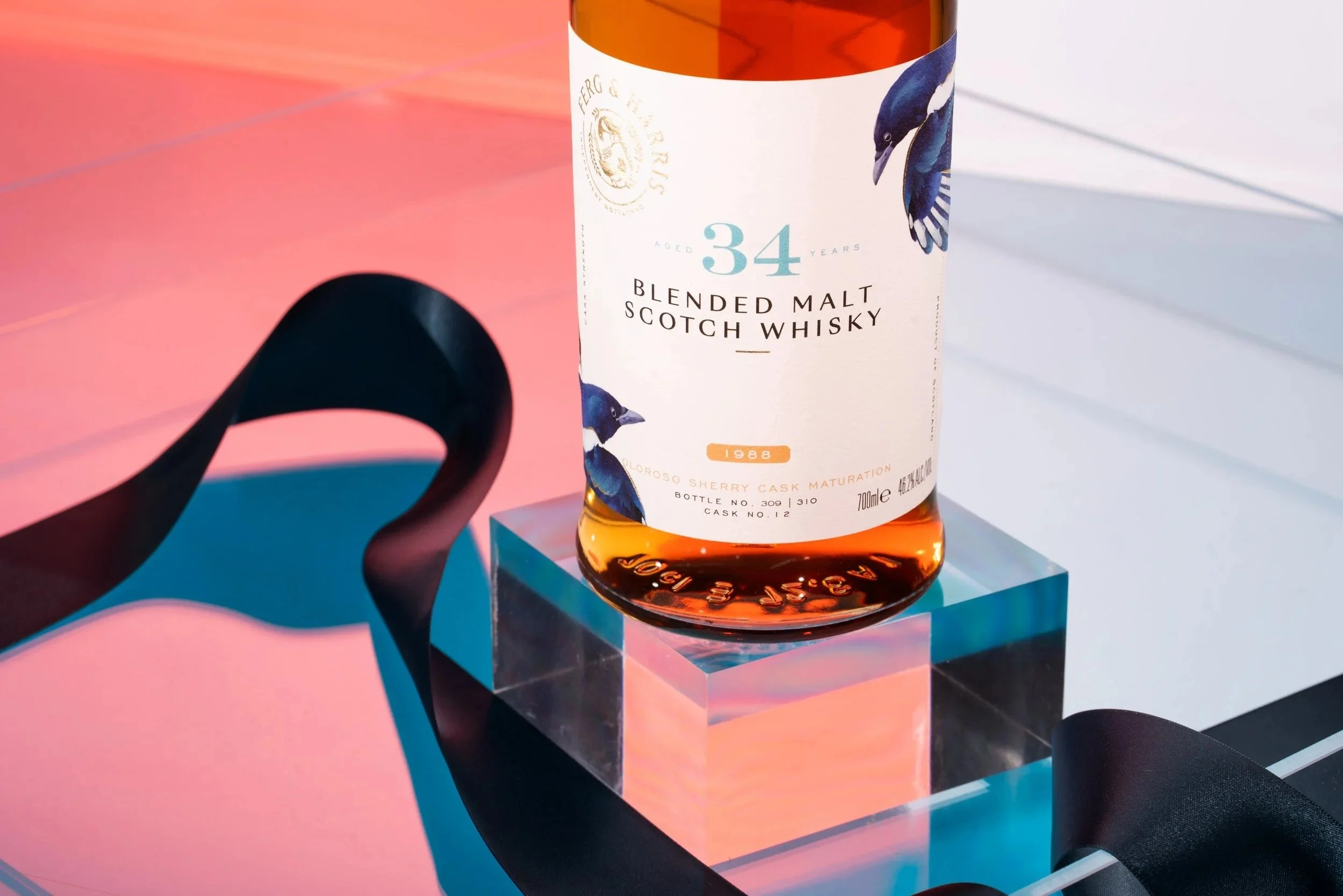 A bottle of blended malt scotch whisky on a clear acrylic block, with colorful lighting and a black ribbon.