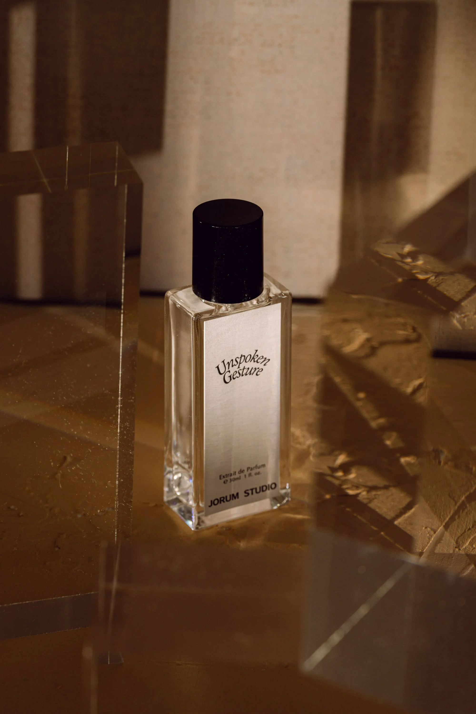 A perfume bottle labeled 'Unspoken Gesture' by Jorum Studio on a reflective surface with decorative gold and metallic accents around it.