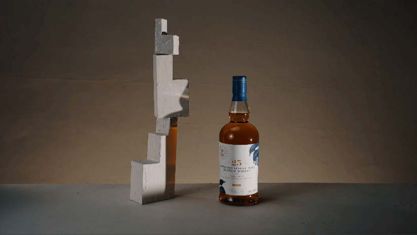 A bottle of whiskey with a blue cap placed on a flat surface next to a tall, abstract sculpture made of stacked small geometrical blocks.