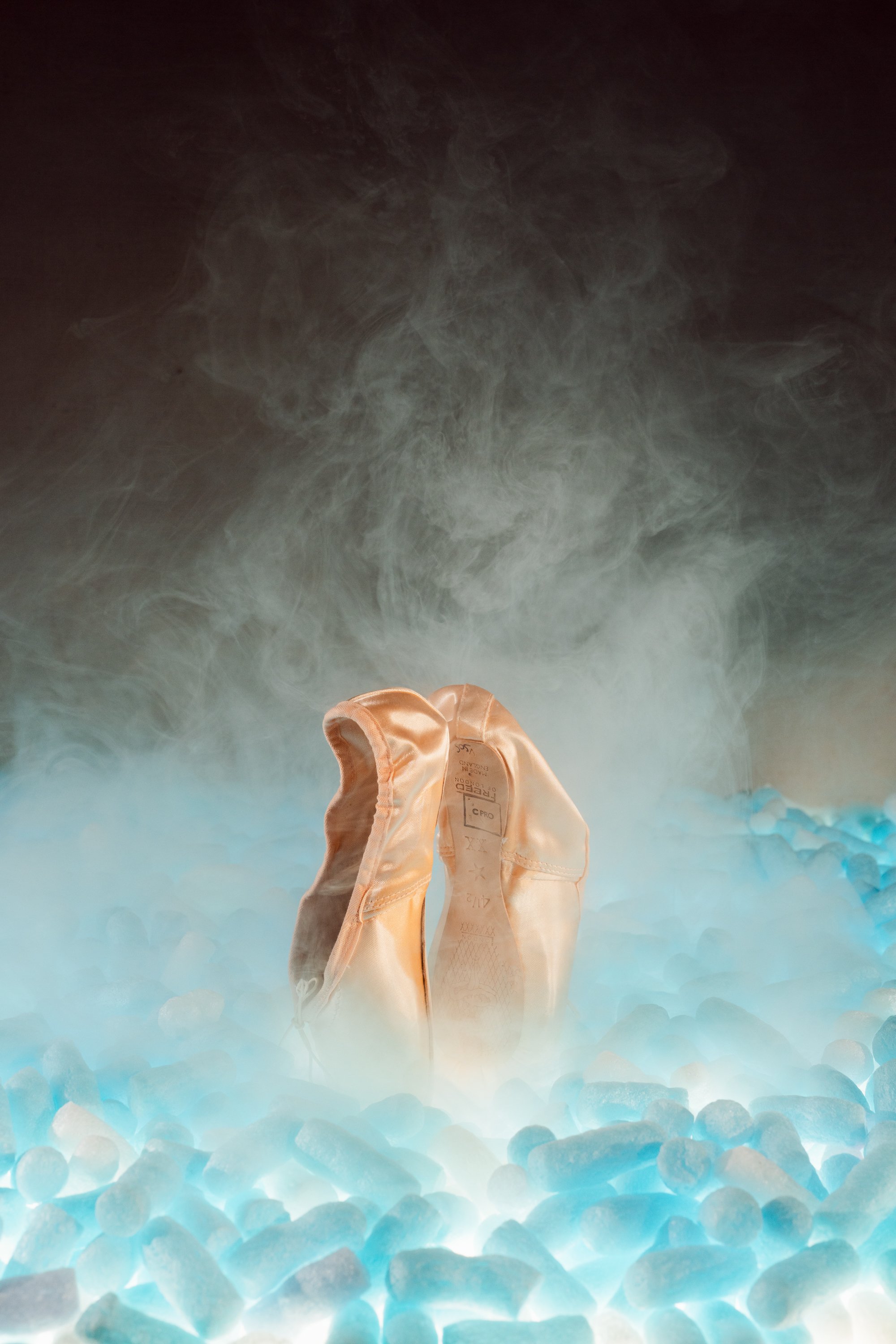 A pair of ballet pointe shoes standing upright on ice with a glowing light underneath, surrounded by mist or fog.