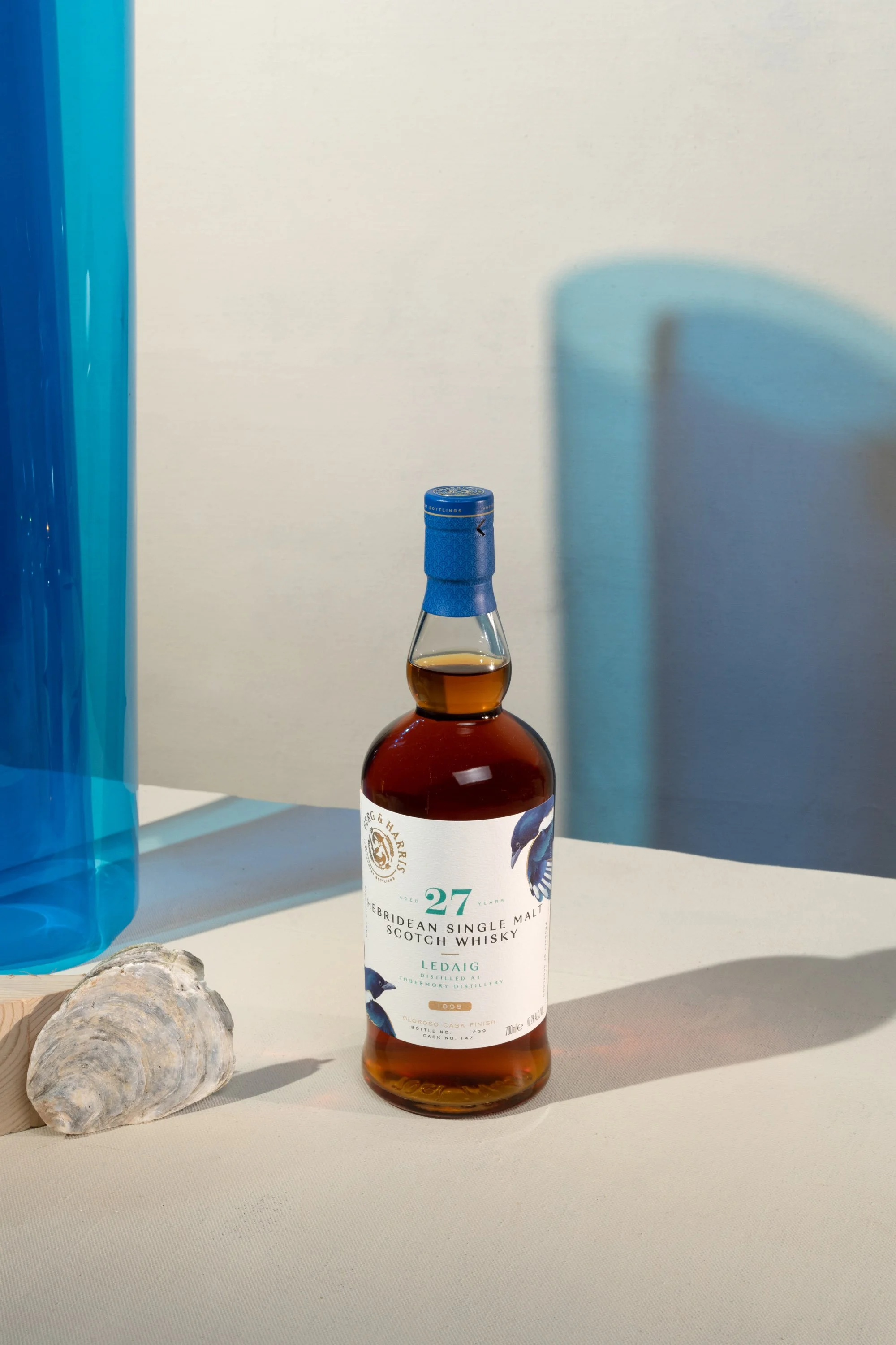 A bottle of Hebridean single malt Scotch whisky on a white surface, with a large shell and a blue translucent object nearby, and a white wall in the background.