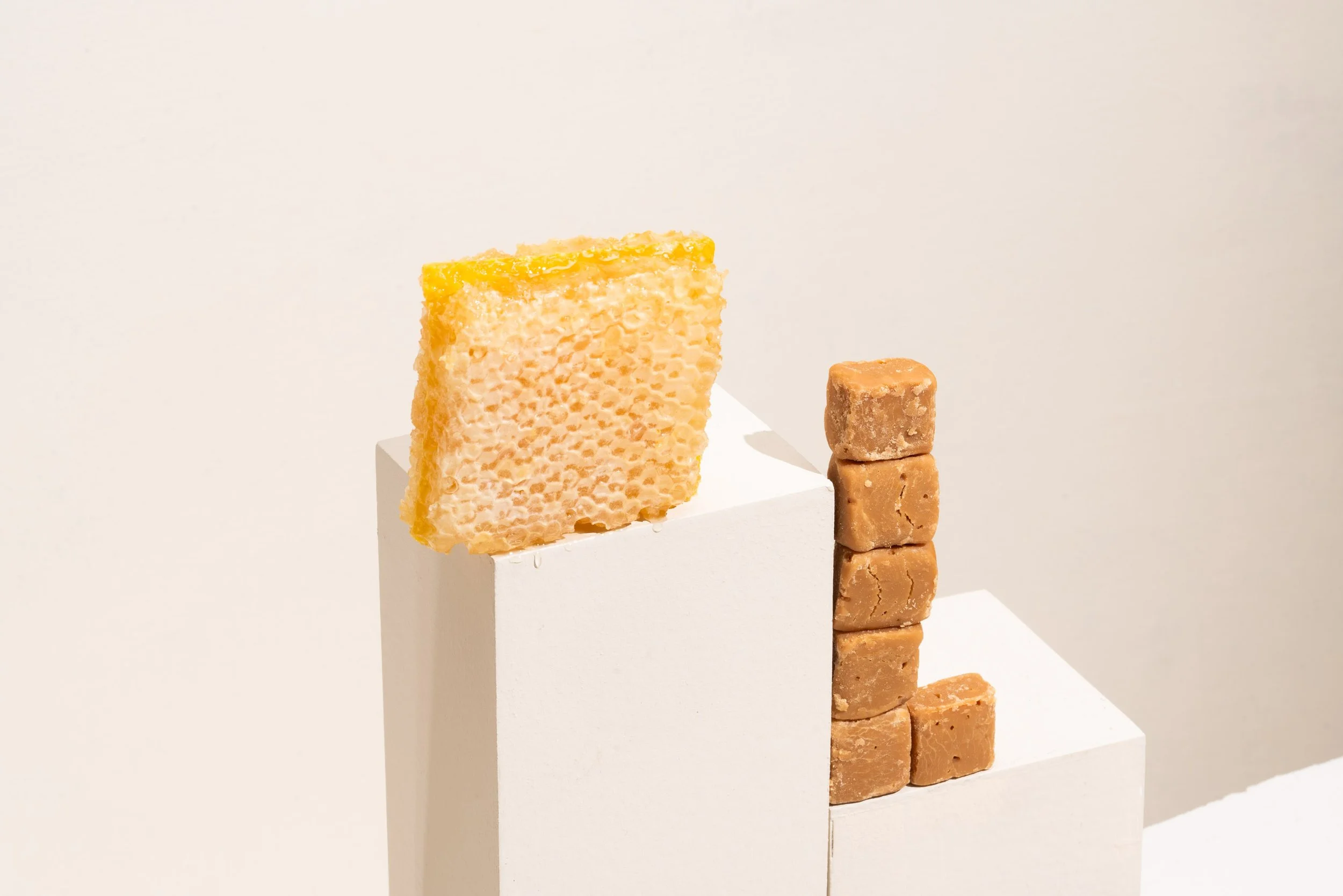 A stack of three beige caramel candies and a block of yellow and white honeycomb soap on a white pedestal against a plain beige background.