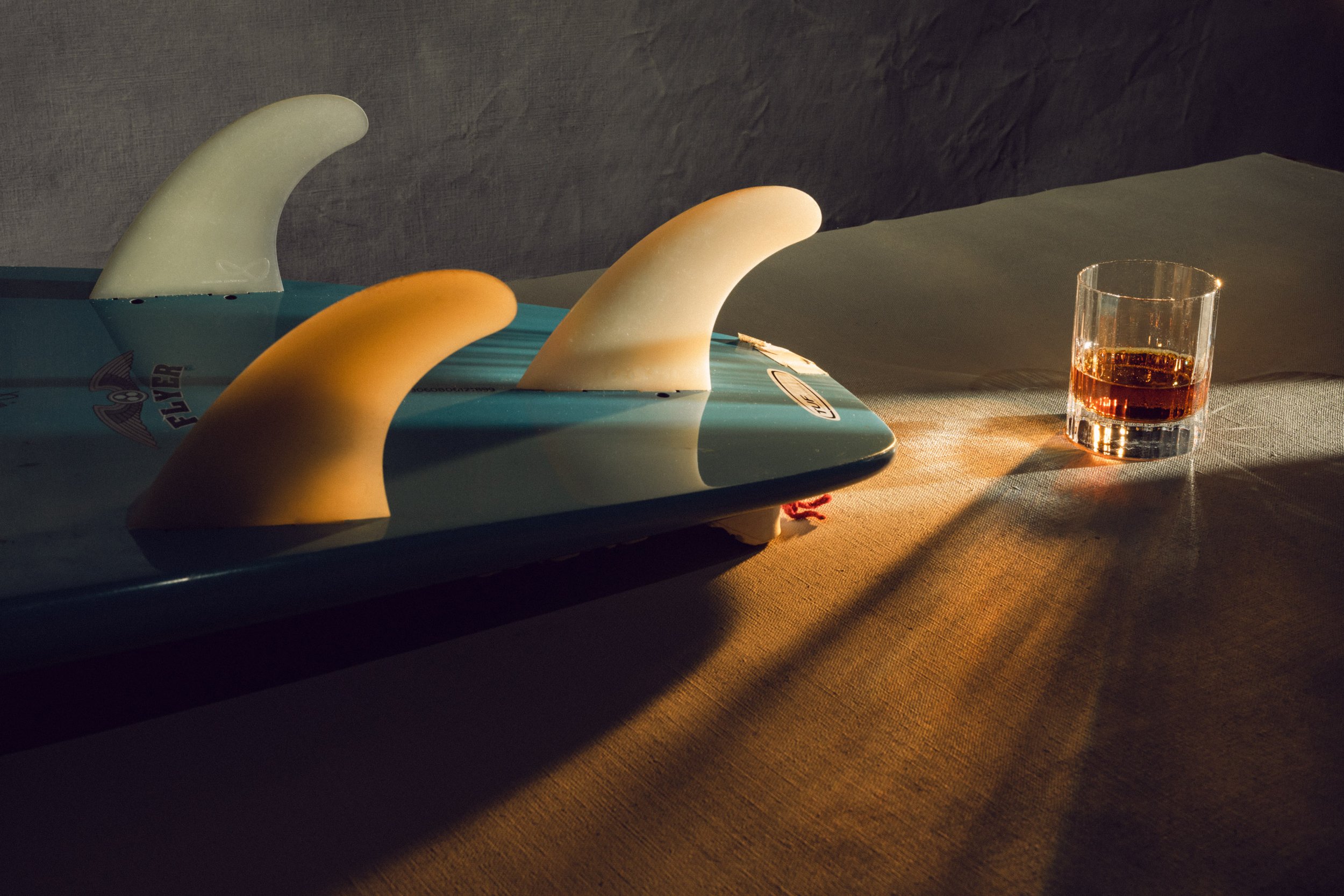 A surfboard with three fins, two white and one yellow, on a table illuminated by warm light, with a glass of dark liquid nearby.