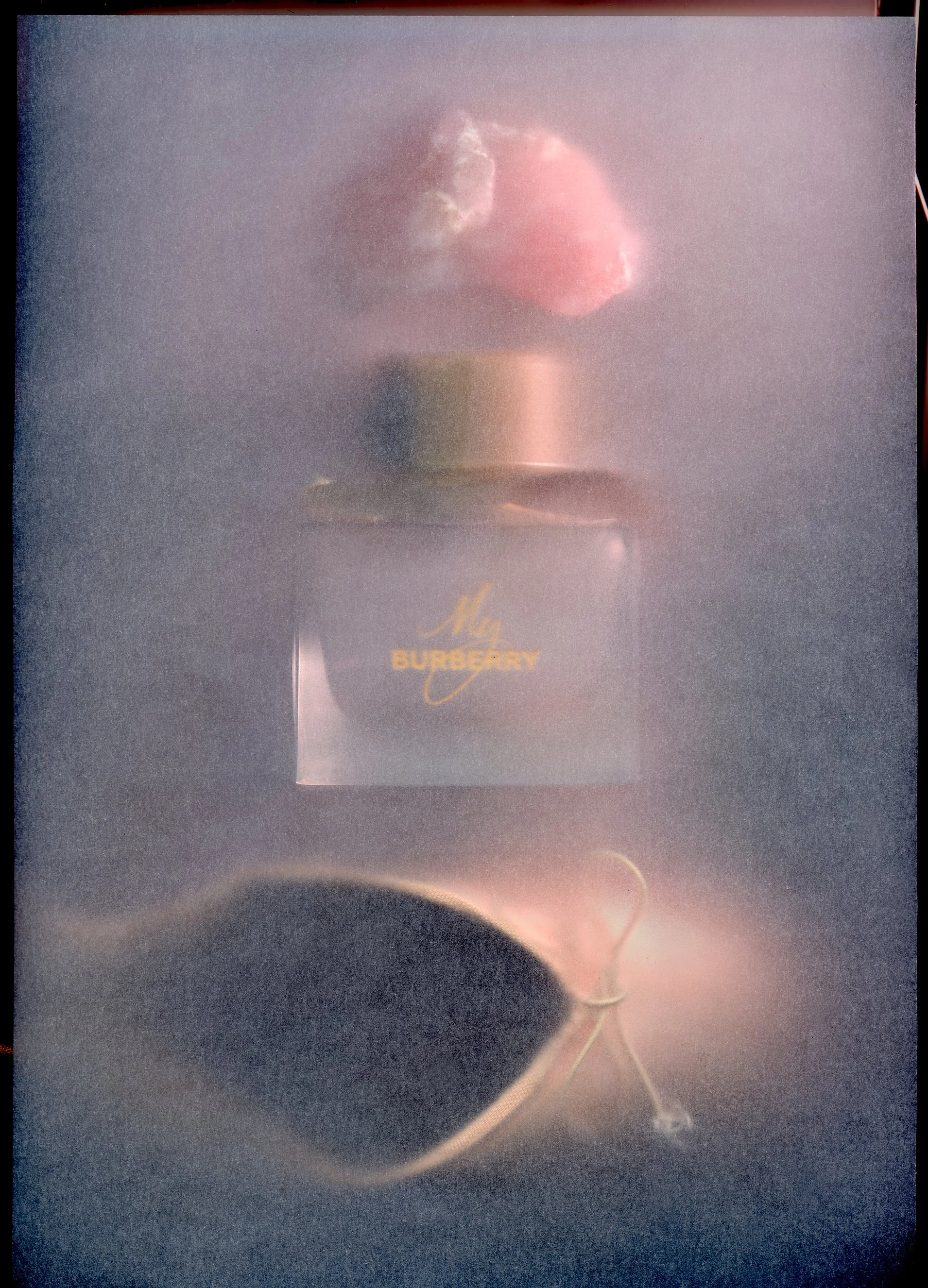 A bottle of Burberry perfume with a pink flower on top and a string with a small bell next to it, against a textured background.
