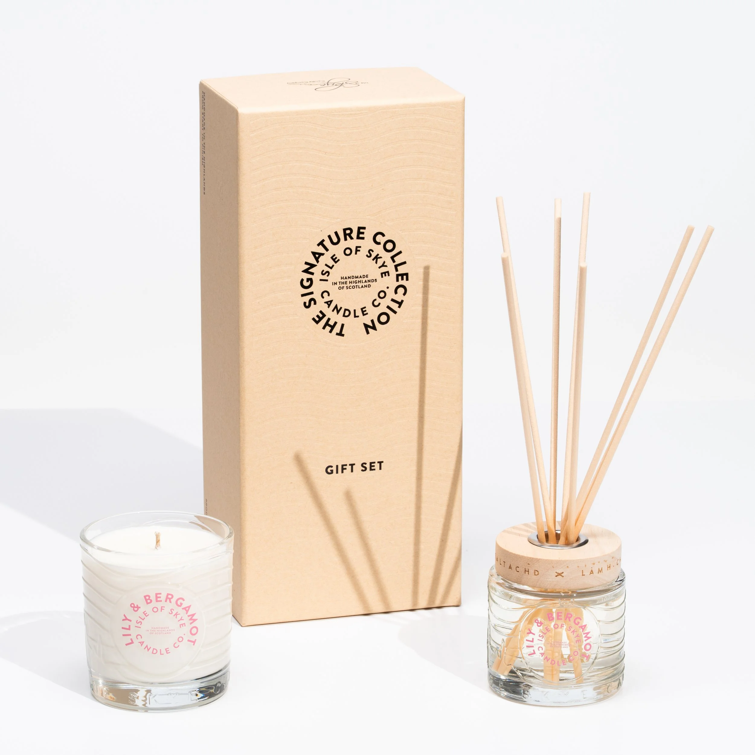 A gift set containing a small scented candle in a glass container with a pink label, a glass jar with wooden reed diffusers with a matching pink label, and a beige cardboard box with black text reading 'The SignaTURE Collection, Isle of Skye, Candle,
