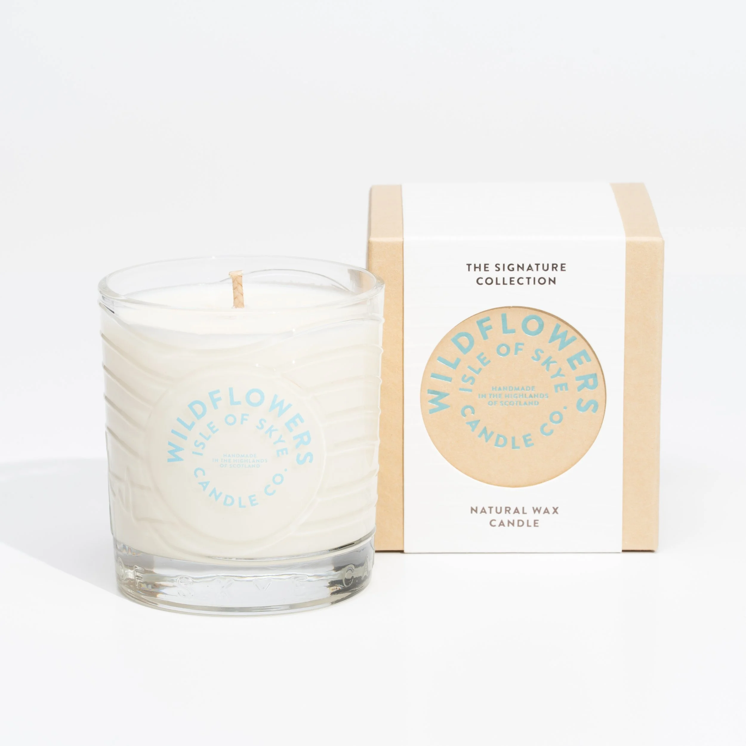A white wax candle in a clear glass holder with a blue and beige 'Wildflowers Candle Co.' label, next to a beige and white box packaging.
