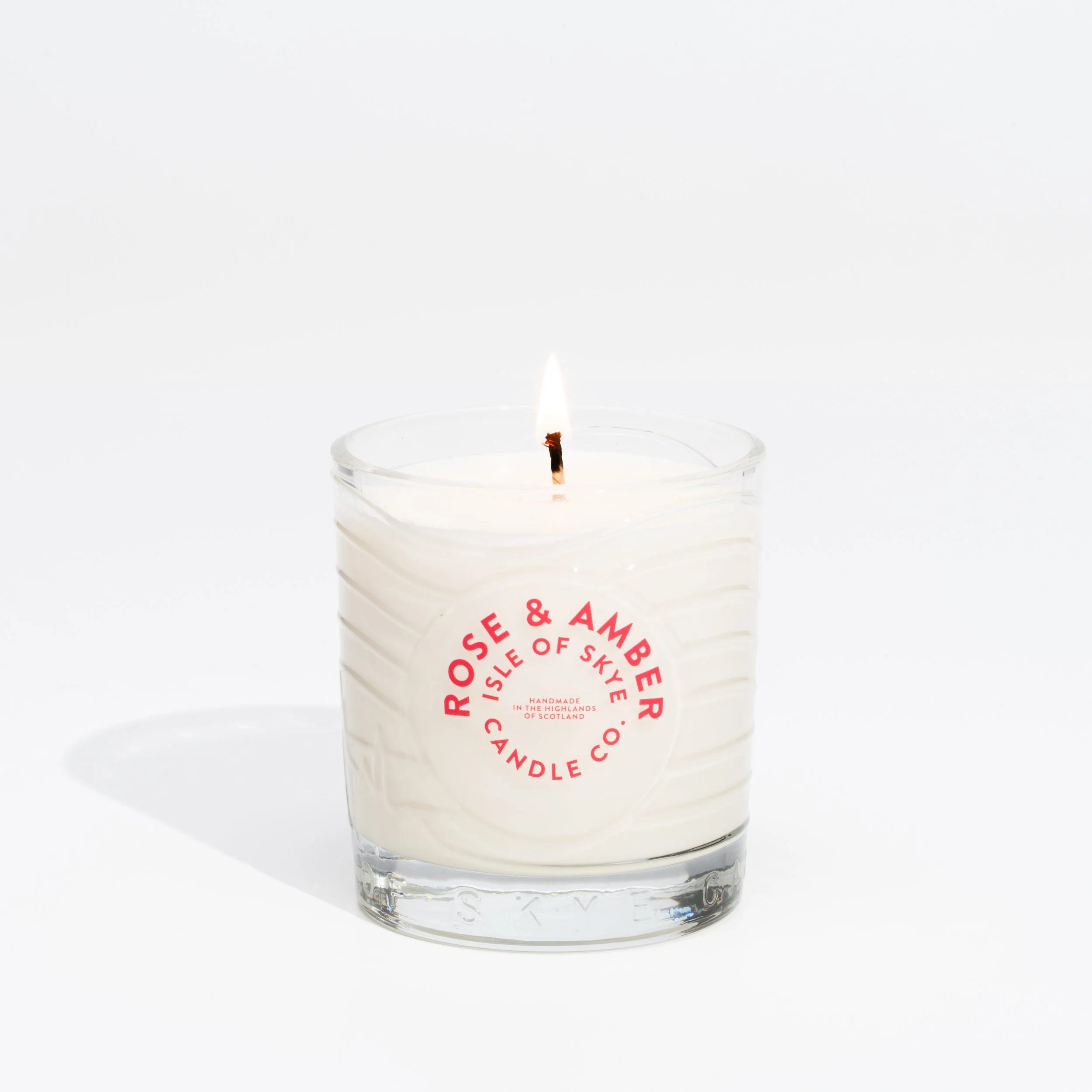White candle in a glass container with a black wick and small flame, label reads 'Rose & Amber Isle of Skye Candle Co.' on a white background.