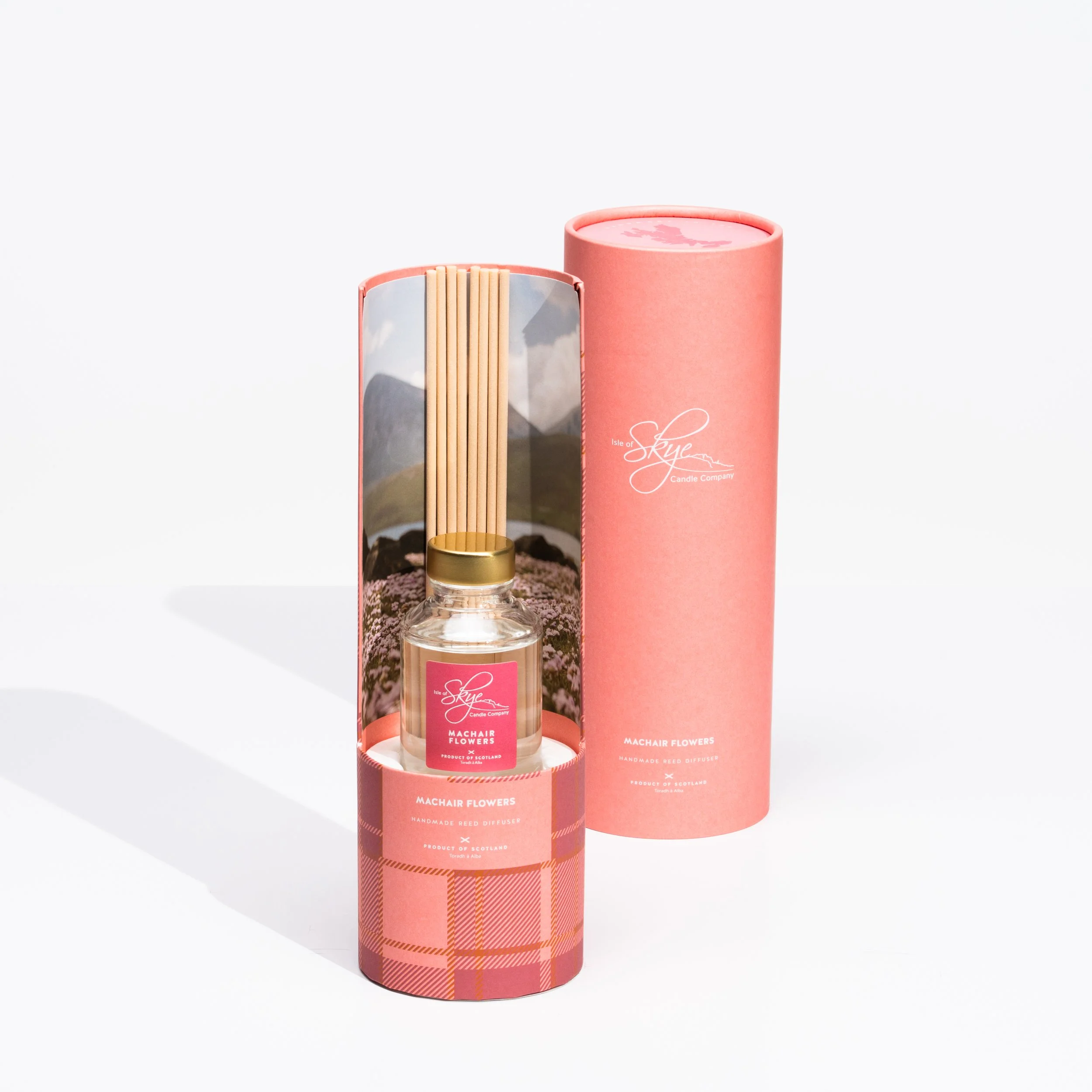 A scented reed diffuser with a glass bottle, light wood reeds, and a pink label, inside a pink cylindrical box with branding, against a white background.