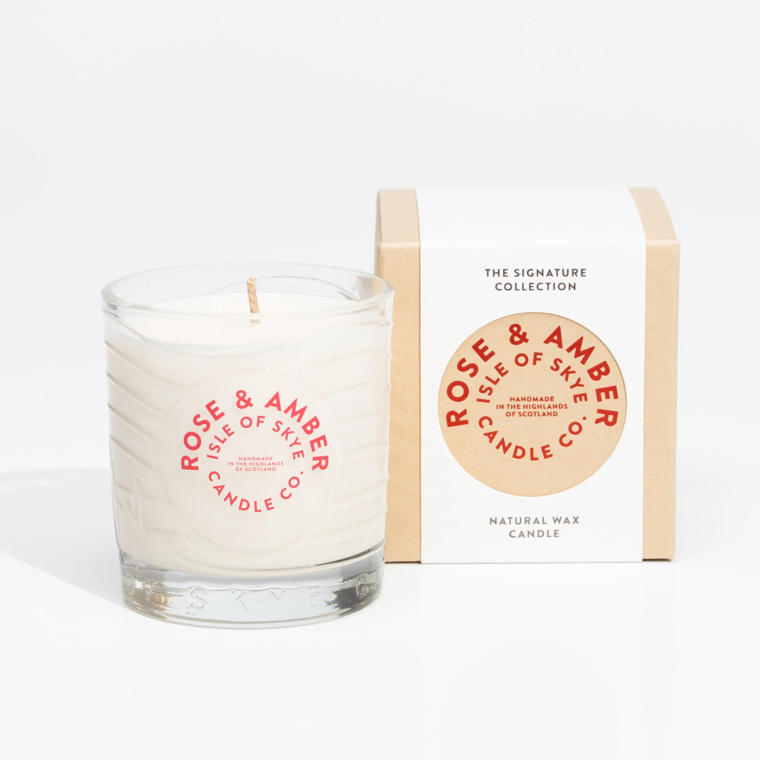 A white scented candle in a glass container with a label reading 'Rose & Amber Isle of Skye Candle Co.' placed beside its beige and white packaging box on a white background.