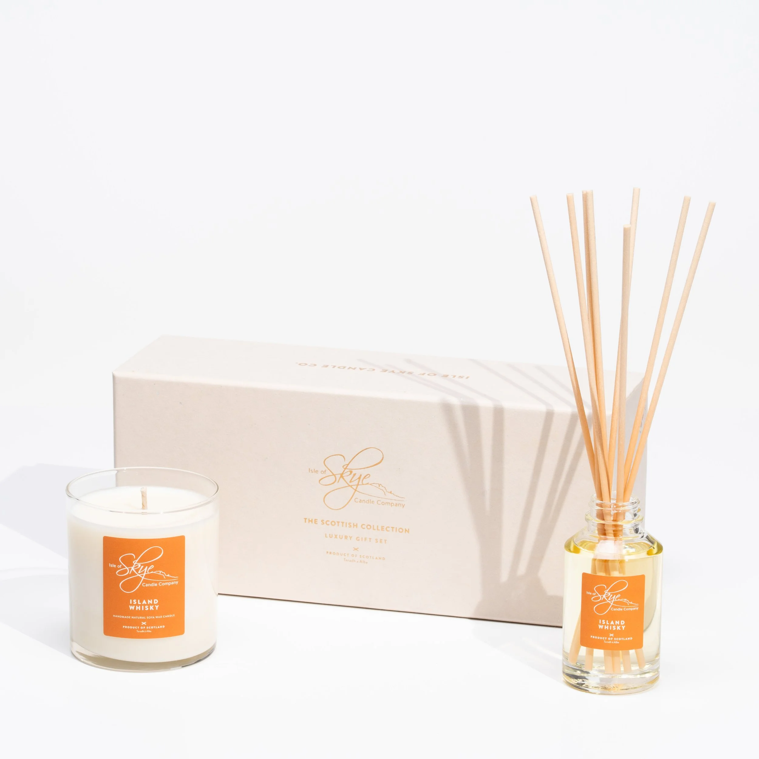 A set of luxury scented candles and reed diffusers from Isle of Skye Candle Company, featuring a white candle in a glass jar with an orange label, a box labeled 'The Scottish Collection,' and a small glass bottle with reed sticks, all on a white back