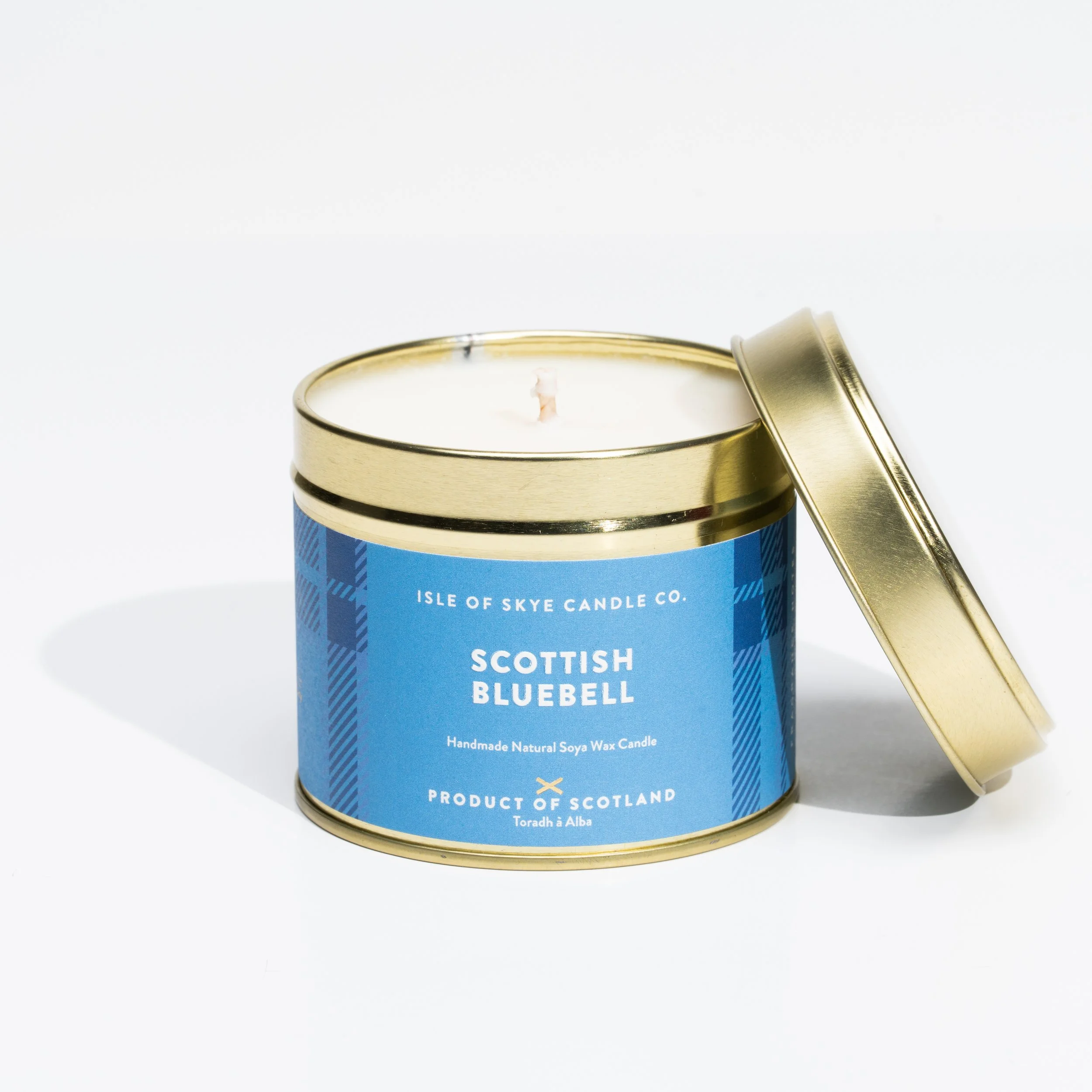 A lit candle in a blue tin container with a gold lid, labeled 'Scottish Bluebell' by Isle of Skye Candle Co., made of natural soya wax, with a white wick and a blue plaid pattern on the label.