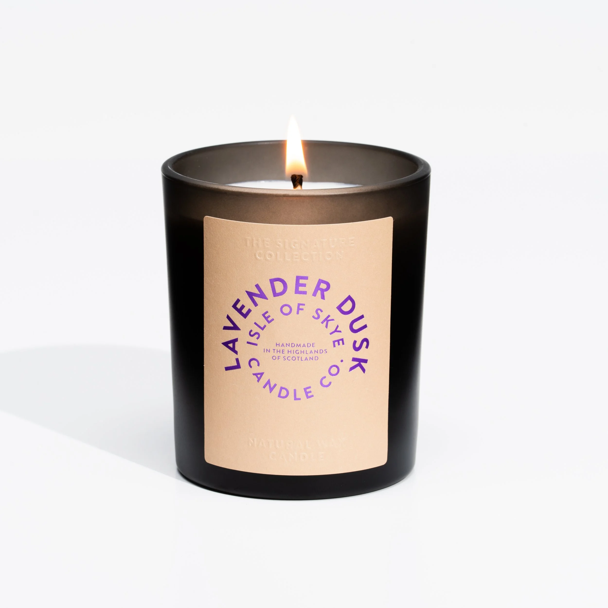 Black candle with a lit flame and a beige label, reading 'Lander Dusk Candle Co., Isle of Skye, Handmade in the Highlands of Scotland, Natural Wax Candle' on a white background.