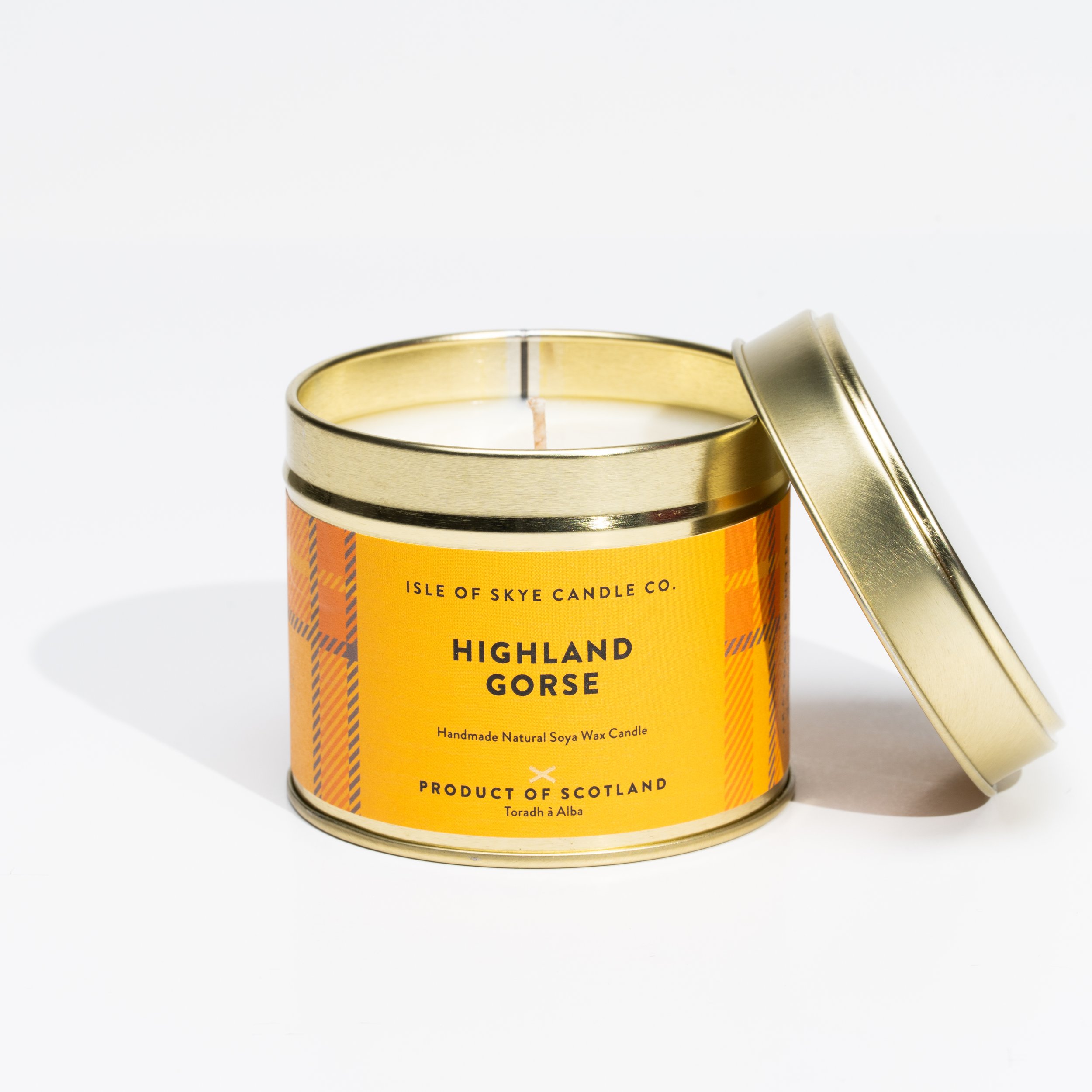A tin container with a golden lid, labeled Highland Gorse scented candle, handmade from Scotland, with a burnt wick inside.