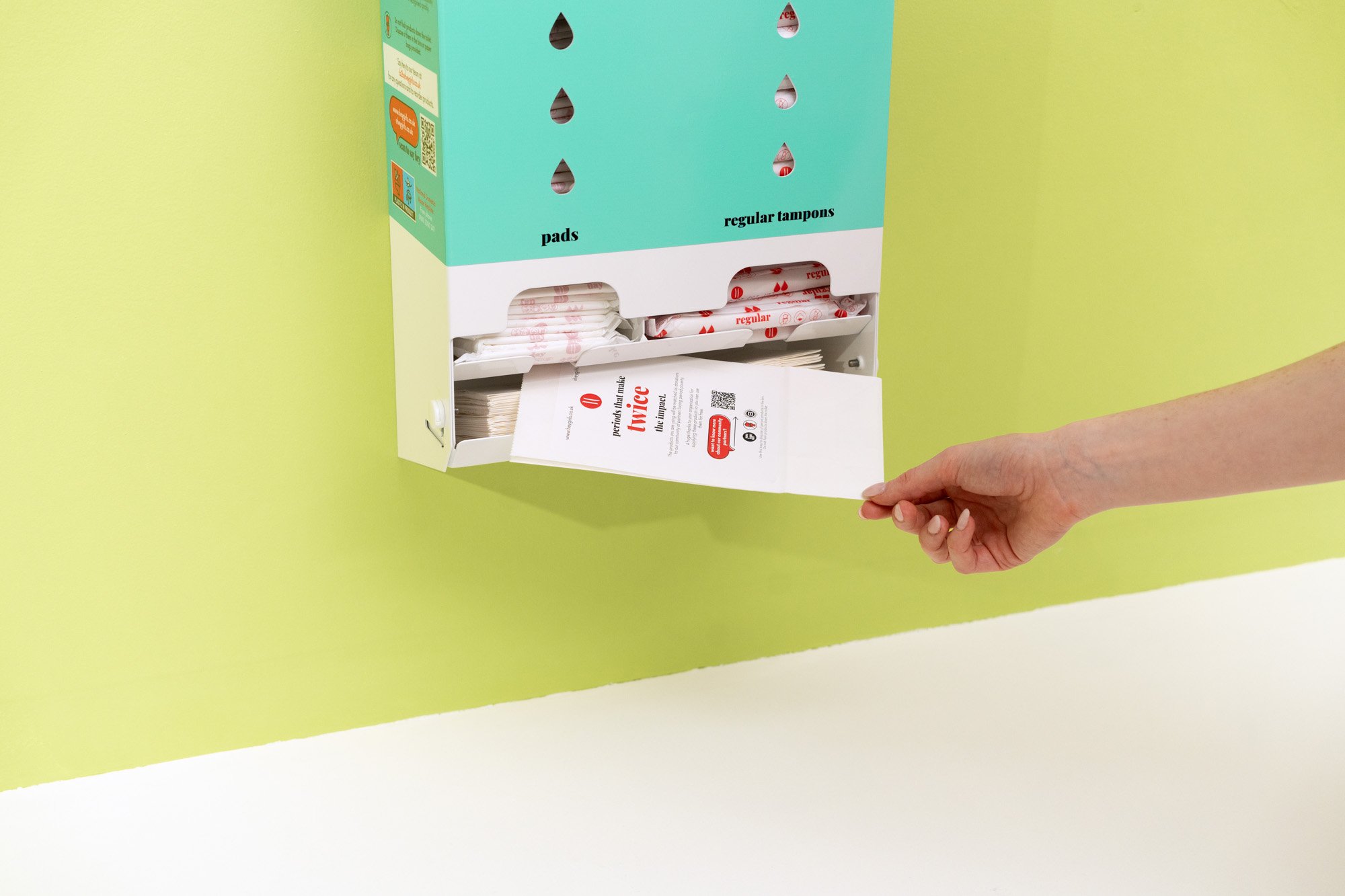 A hand reaching into a teal and white wall-mounted box containing menstrual pads and regular tampons.