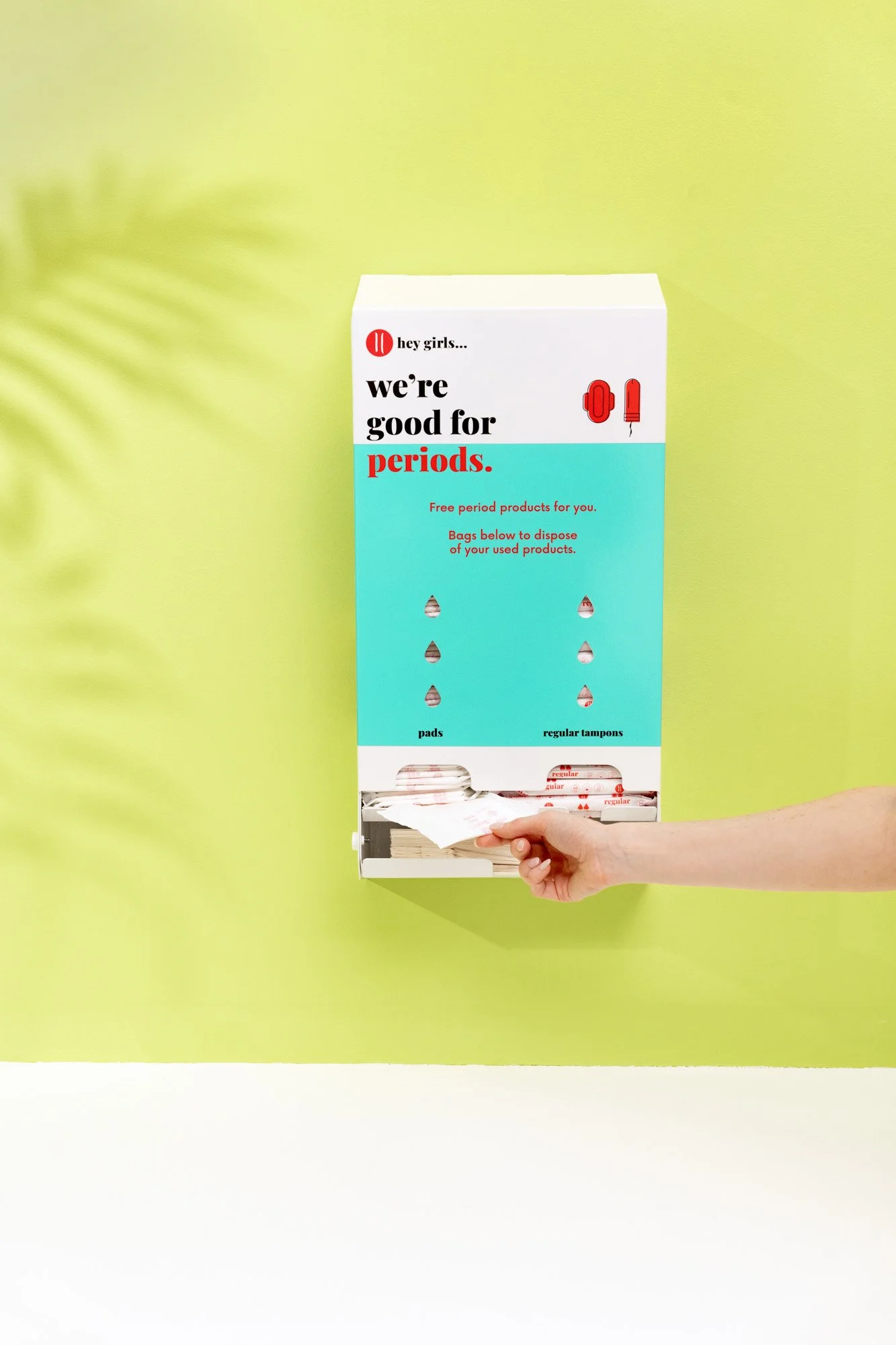 A teal and white dispenser with the text 'hey girls... we're good for periods.' offering free period supplies, with a hand placing a pad into a bag.