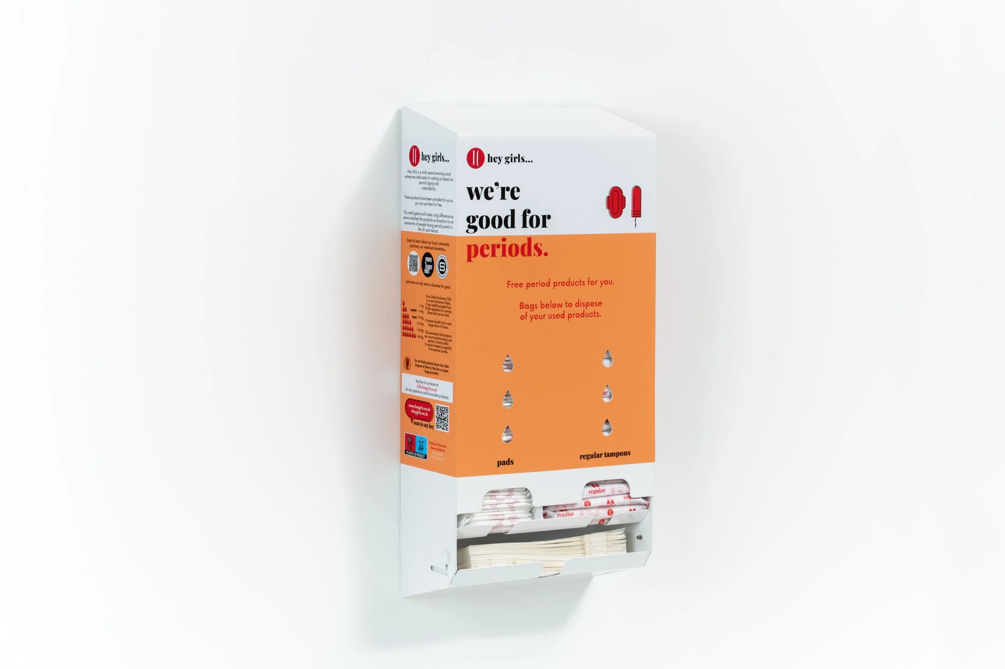 A wall-mounted dispenser with a transparent bottom section holding sanitary pads and tampons. The dispenser is white with an orange label that reads 'hey girls... we're good for periods' and includes instructions and icons for menstrual products.