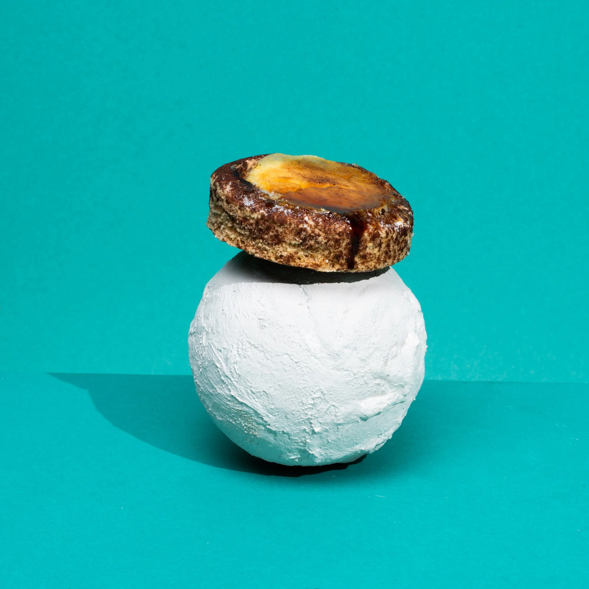 A cinnamon roll with icing on top, placed on a black plate, balanced on a white spherical object, against a blue gradient background.