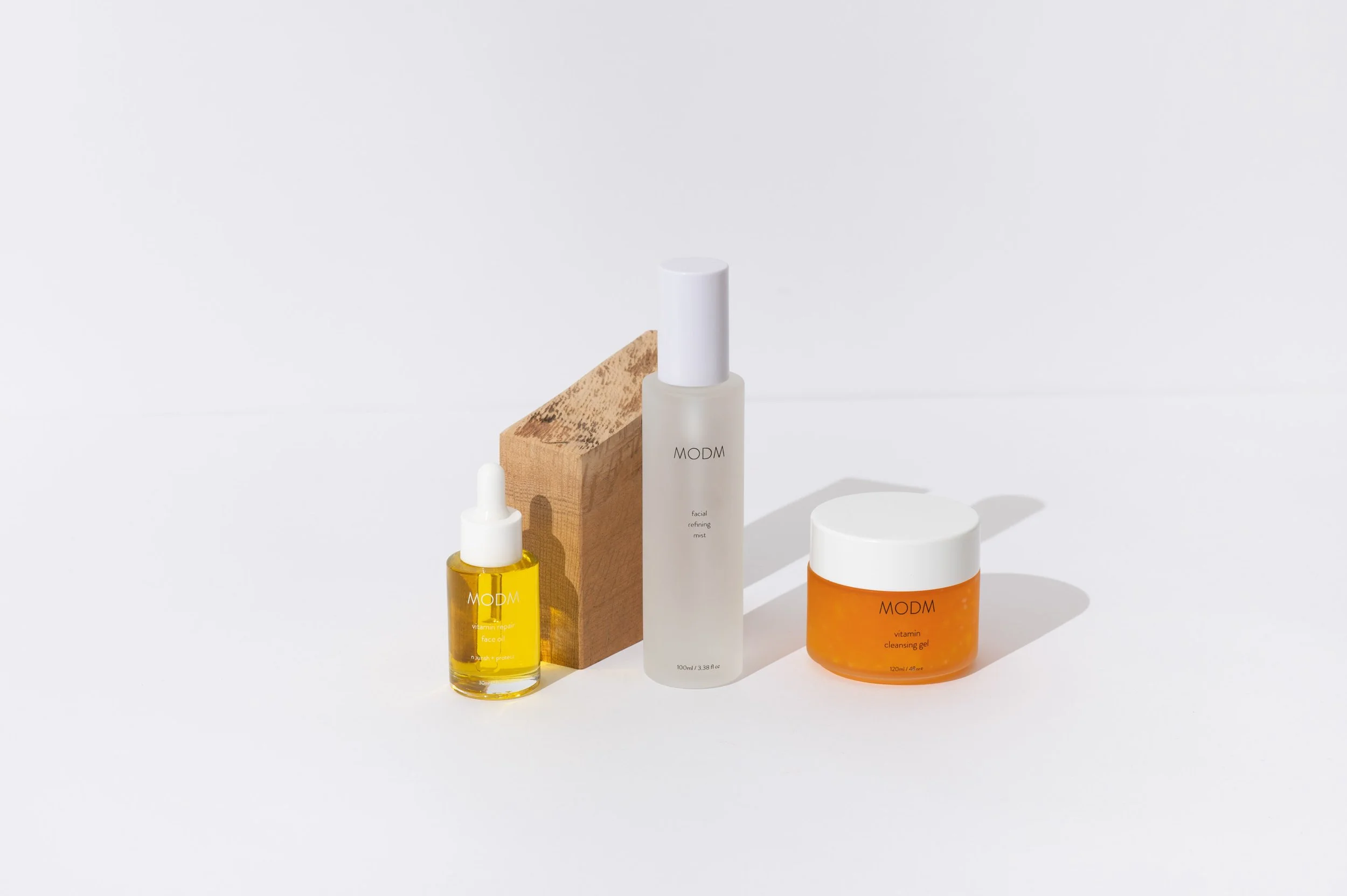 Collection of three skincare products including a yellow face oil with dropper, a white facial mist in a frosted bottle, and an orange cleansing gel in a jar, arranged on a wooden block against a white background.