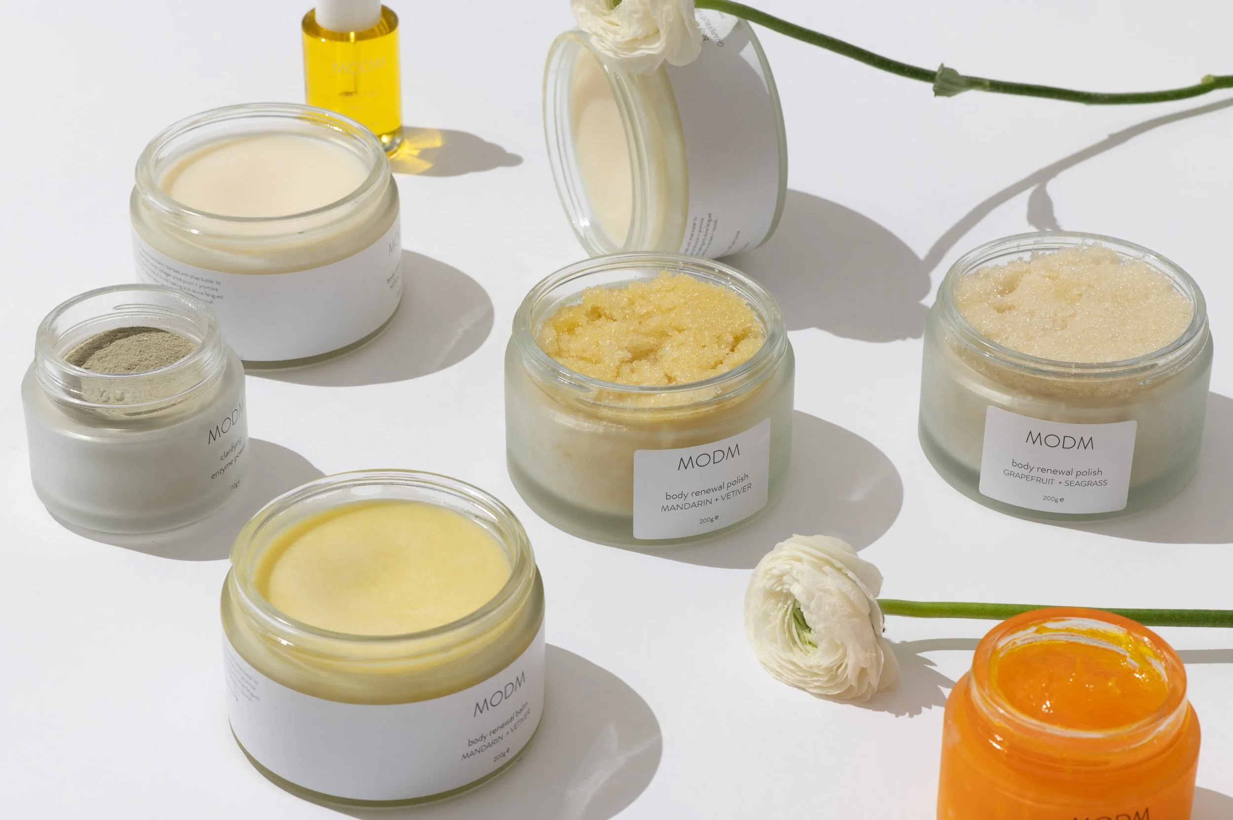 Assorted skincare products in jars and bottles on a white surface with white and orange flowers.