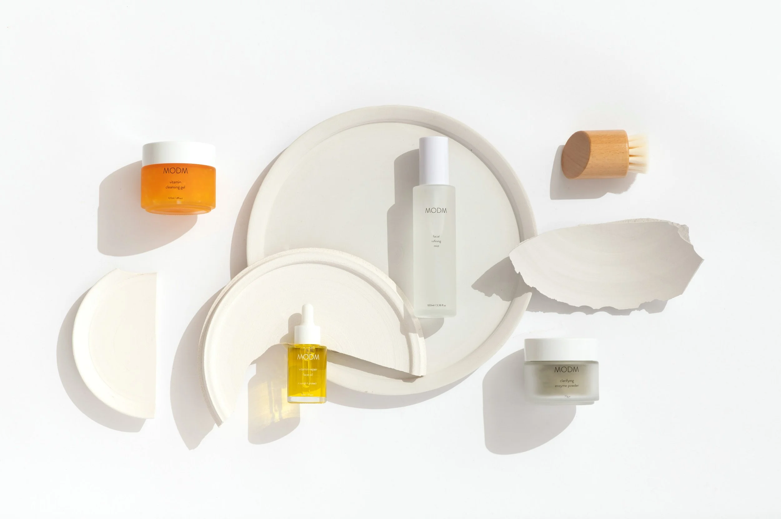 Skincare products on white circular trays, including serums, creams, a bamboo brush, and shell-shaped objects, arranged on a white background.