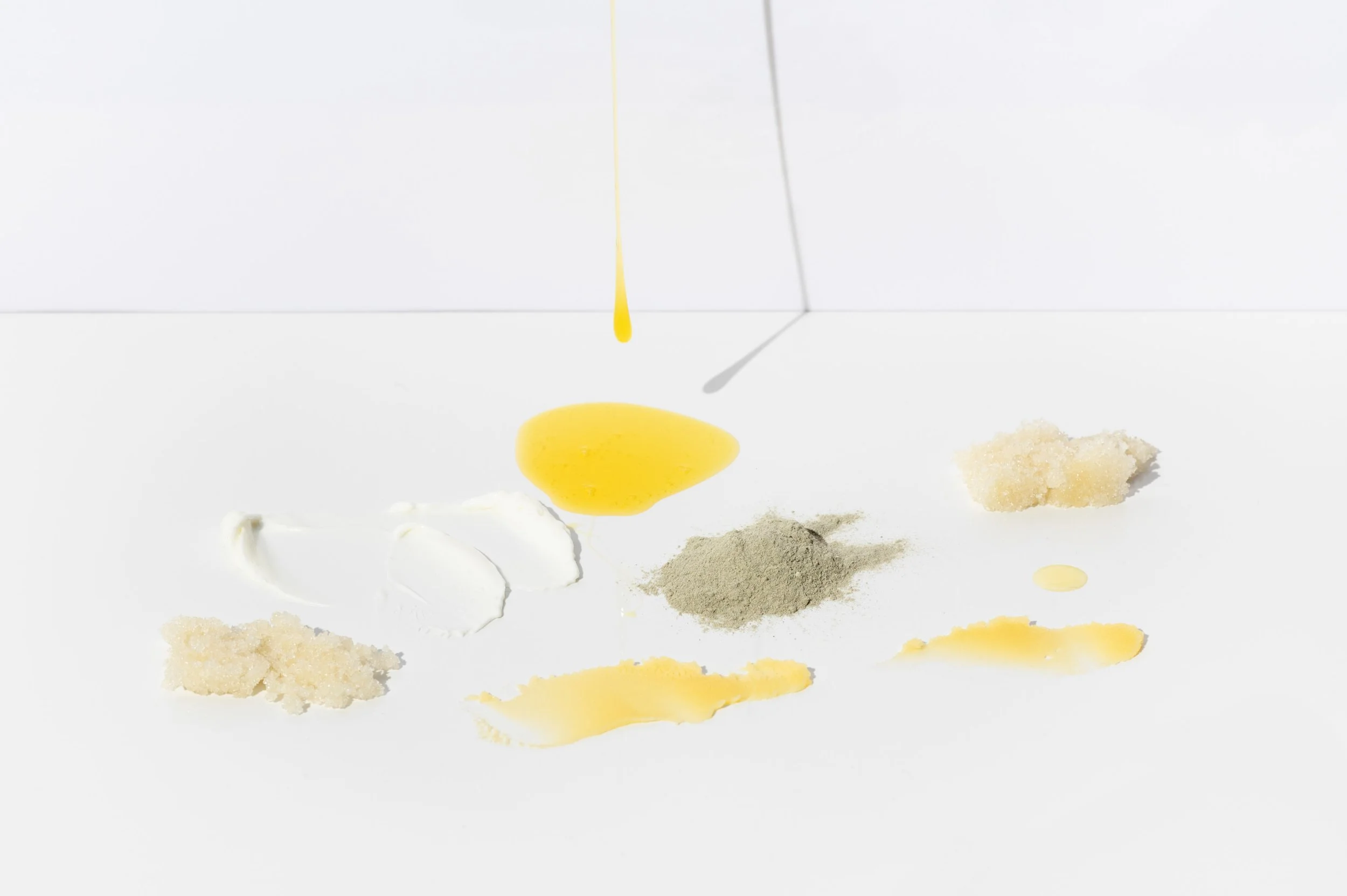 Yellow egg yolk dripping from above onto a white surface with various food ingredients including sugar, flour, and liquids arranged beneath it.
