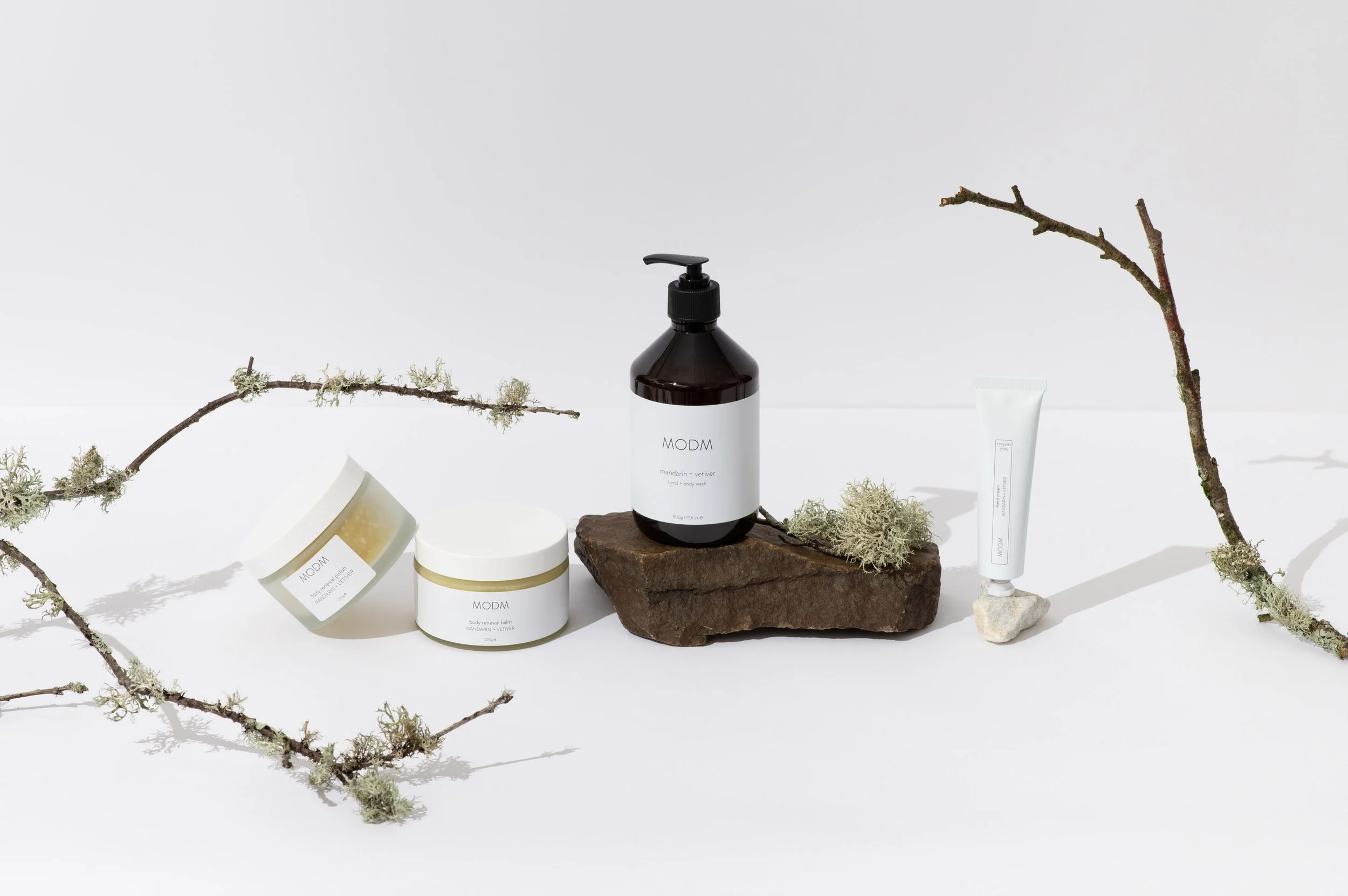 Collection of skincare and body care products, including jars and a pump bottle, arranged on a white background with moss and branches for decoration.