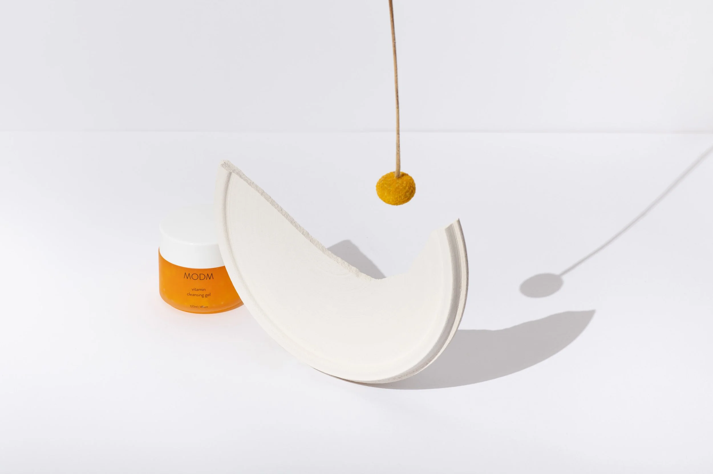 A white half-sphere object with a yellow sphere on a stick above it, casting a shadow on a white background. There is an orange container labeled MODM Vitamin Cleansing Gel nearby.