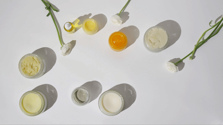 Multiple jars and bottles of skincare products on a white surface with some green flower stems.