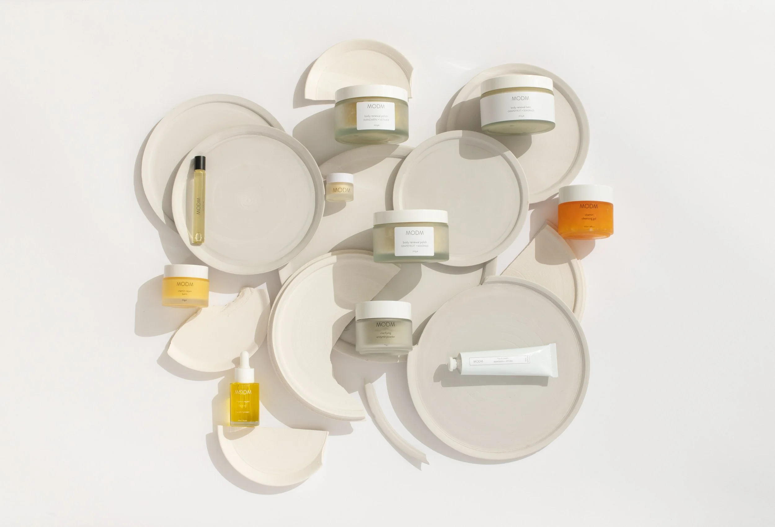 Arrangement of skincare products on white and beige circular trays and plates, including jars, bottles, and tubes in white, yellow, orange, and clear packaging.